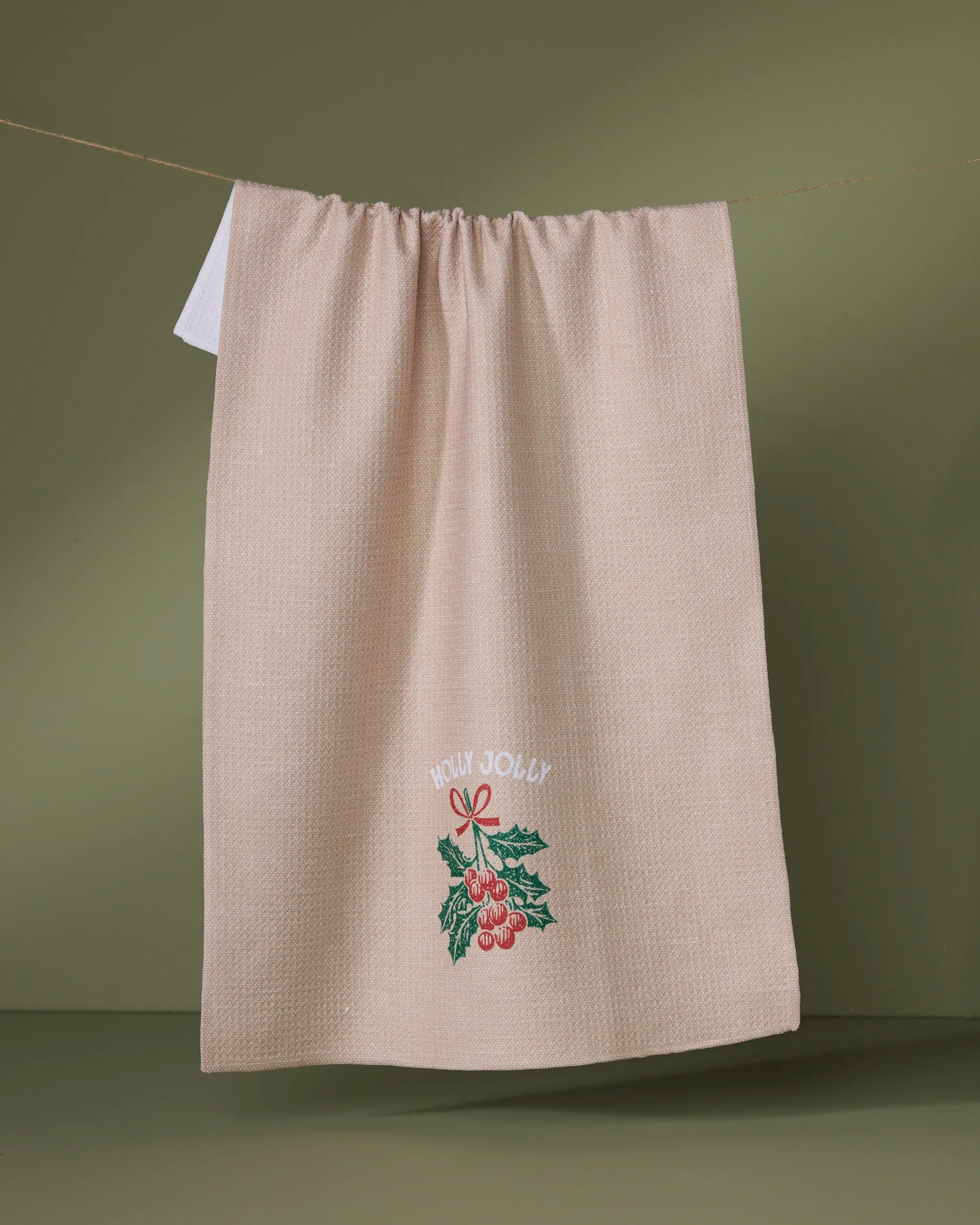 The Geometry Merryberry kitchen towel, crafted from recycled materials, hangs on a string against a green background. It features embroidered red berries, green leaves, and "HOLLY JOLLY" in white above the design for eco-friendly charm.