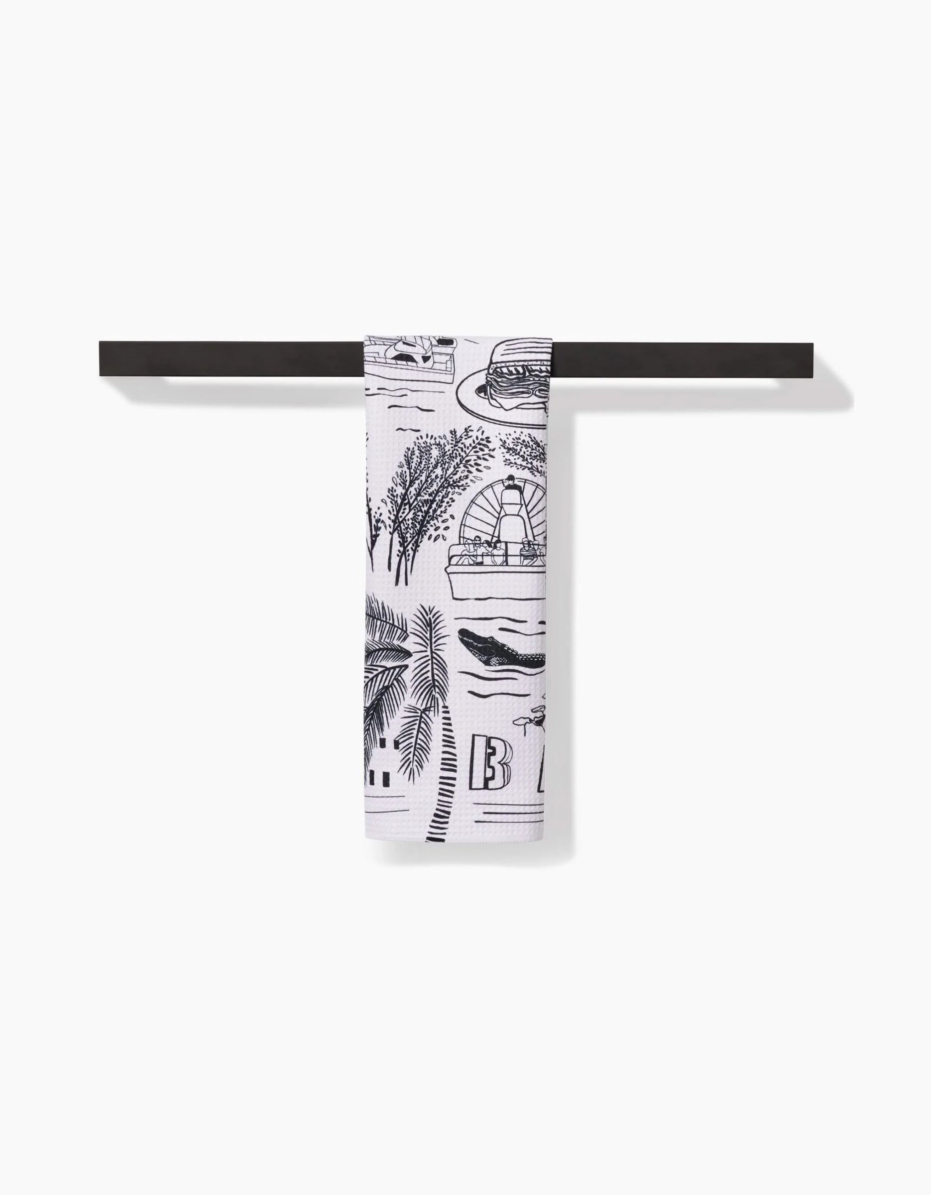 The Miami tea towel by Elisabeth Wing, featuring black beach-themed illustrations on white and made from recycled materials, hangs neatly from a horizontal black towel bar against a plain white background.