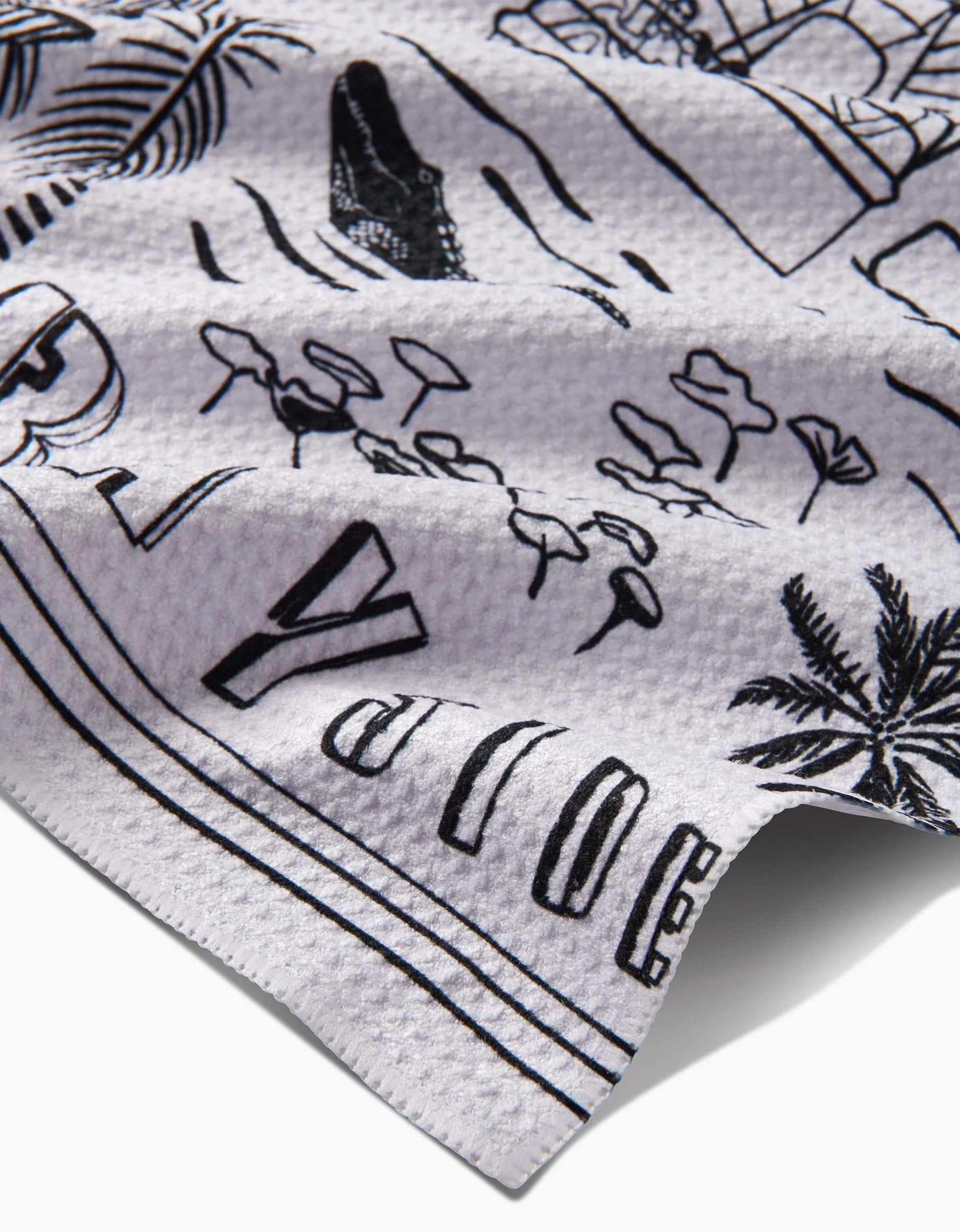 A close-up of the textured white Miami Tea Towel by Elisabeth Wing, showing black line art of palm trees, umbrellas, and an alligator on recycled fabric with bold partial lettering along the edge.