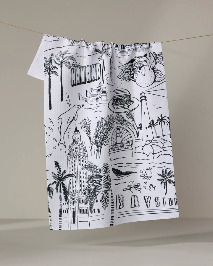 The Elisabeth Wing "Miami" kitchen towel is white, made from recycled materials, and features black line drawings of Miami landmarks, palm trees, a burger, cocktail, and the words HAVANA and BAYSIDE.
