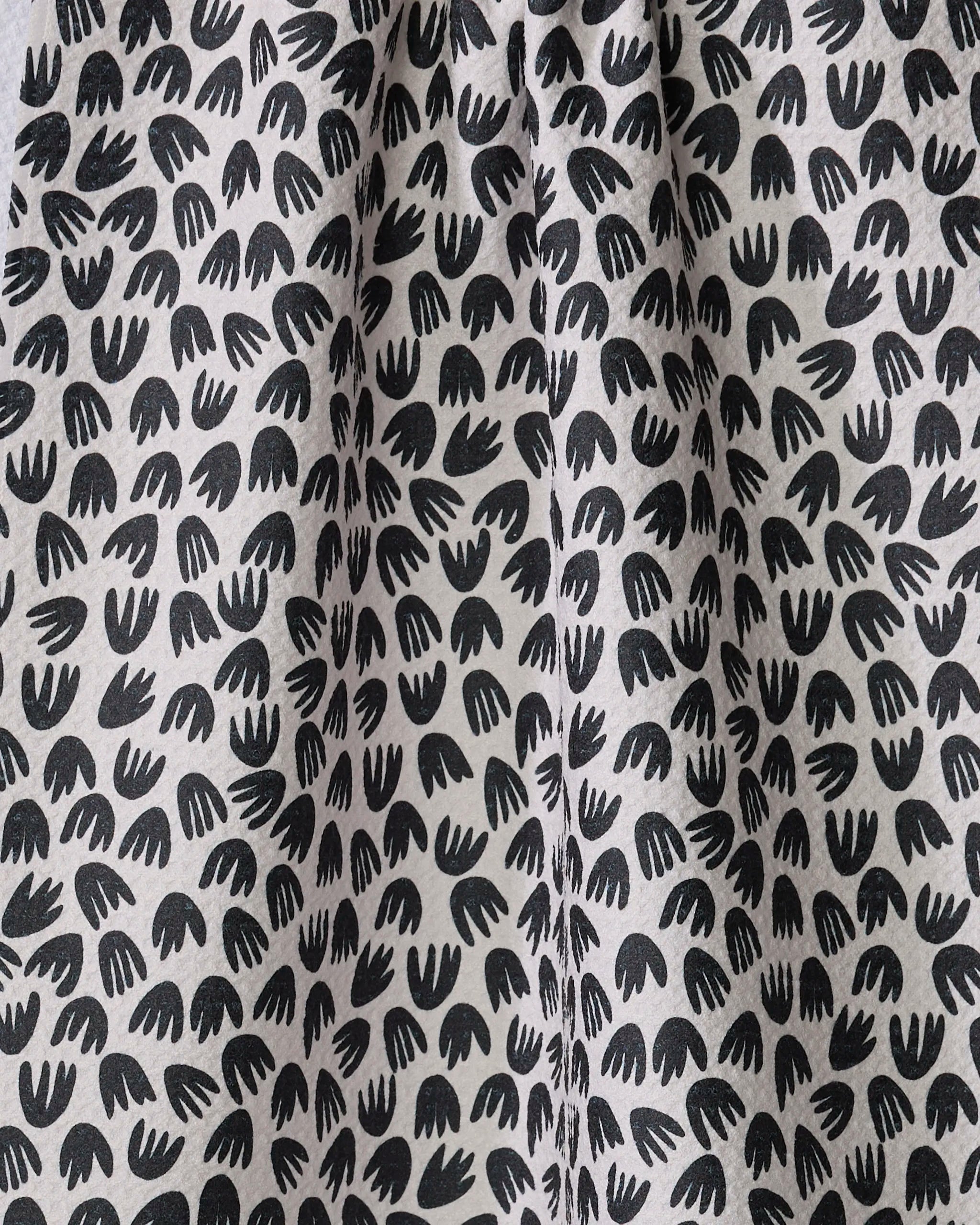 Black abstract bird-footprint shapes are scattered across the white recycled Microbloom kitchen towel by Kayla Ann, forming a repeating pattern on gently pleated fabric with subtle folds and soft shadows.