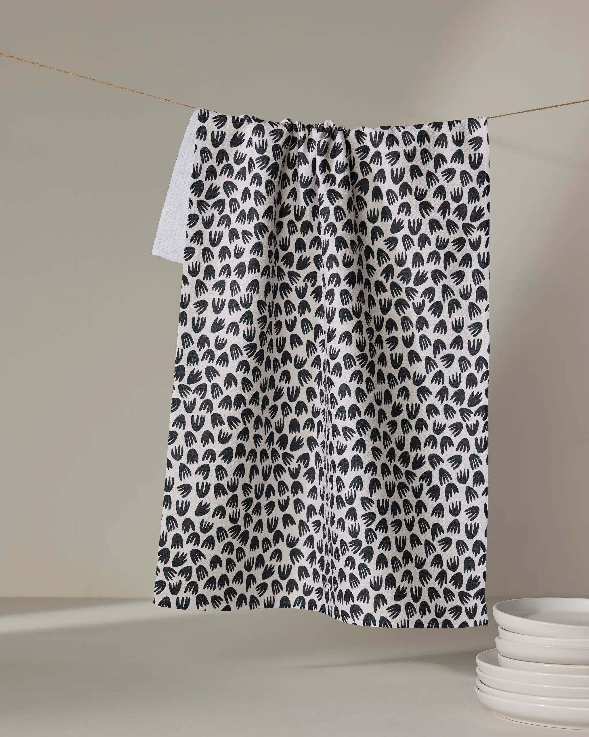 The Kayla Ann Microbloom tea towel, featuring a black handprint pattern, hangs on a clothesline beside stacks of white plates on a neutral surface.