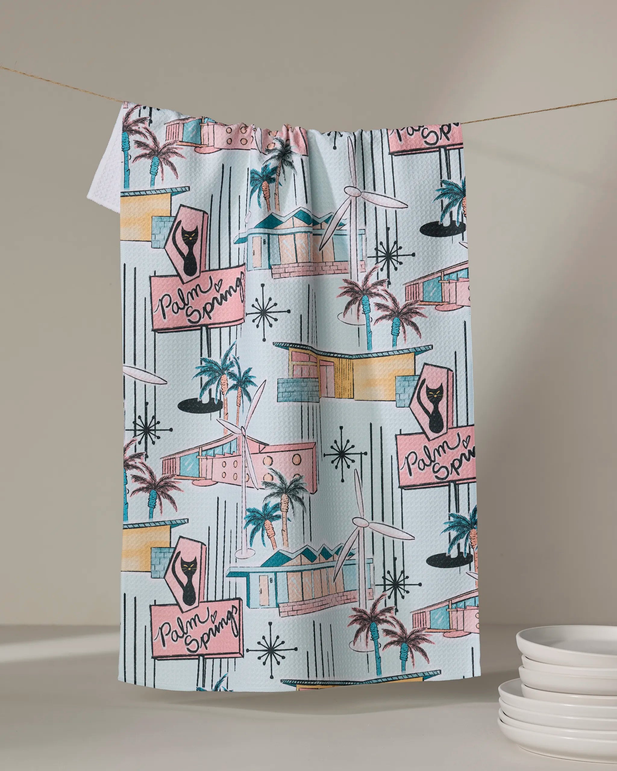 The "Mid Century" tea towel by Julianne Haness features a retro Palm Springs design with pastel houses, palm trees, and pink signs. Quick-drying and made from recycled materials for eco-friendly charm—perfect next to neatly stacked plates.