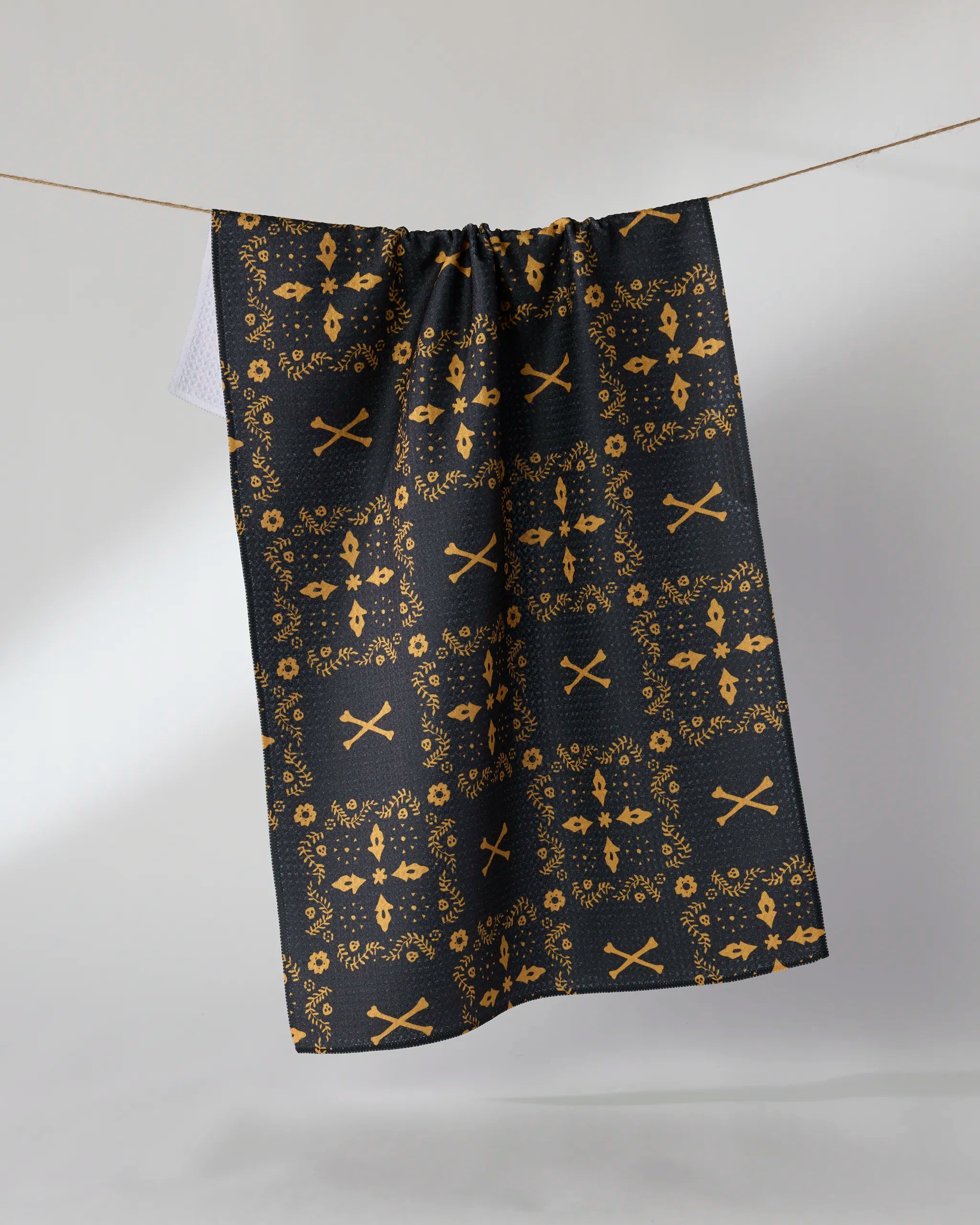 The Geometry Midnight Bandana tea towel, in black with an orange floral and crossed-swords design, hangs from a string against a plain white background.
