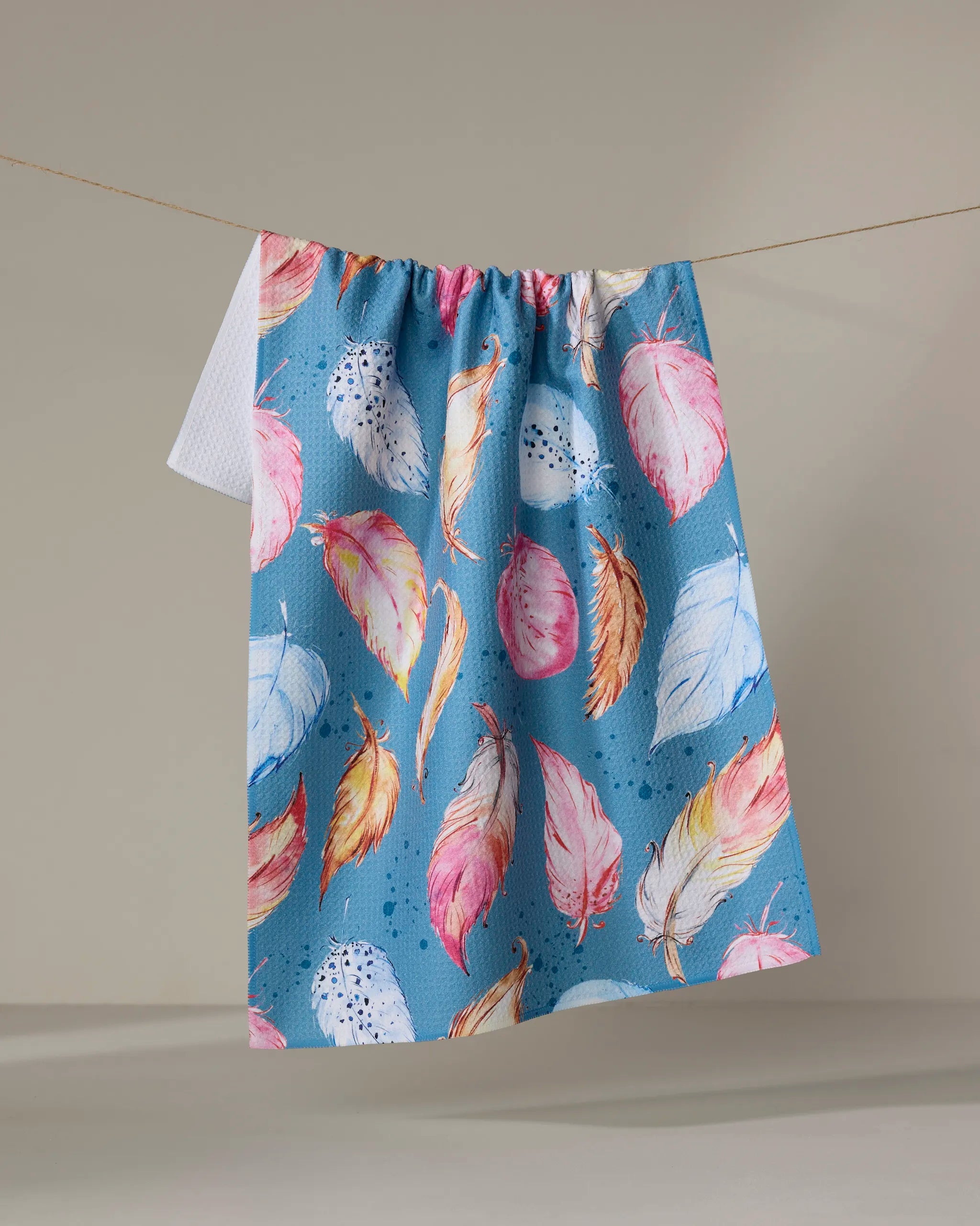 The Elena Fay "Midnight Breeze" tea towel, made from recycled materials, showcases a vibrant feather pattern in pink, yellow, orange, and white and hangs on a clothesline against a plain neutral background.