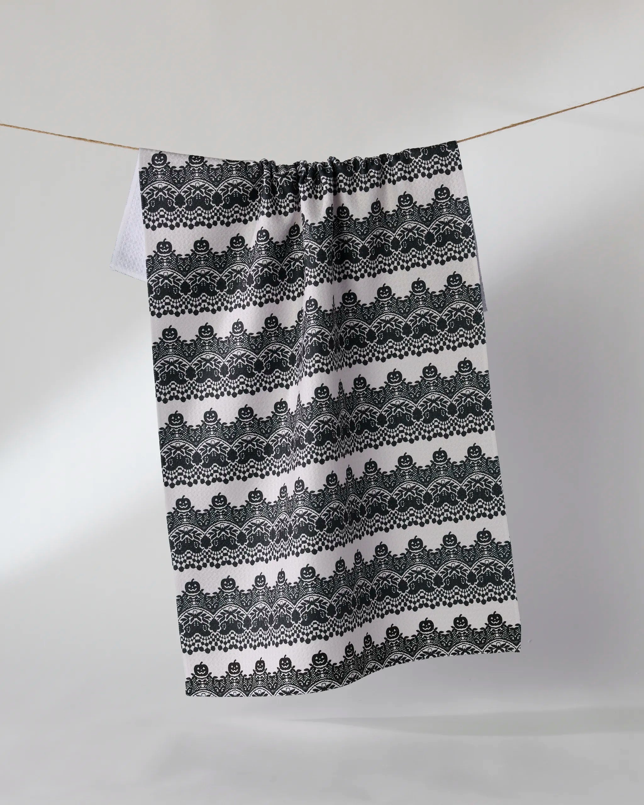 The Geometry “Midnight Lace” kitchen towel, made from recycled materials, hangs on a clothesline against a light gray background. It displays black and white horizontal rows of intricate, symmetrical patterns.
