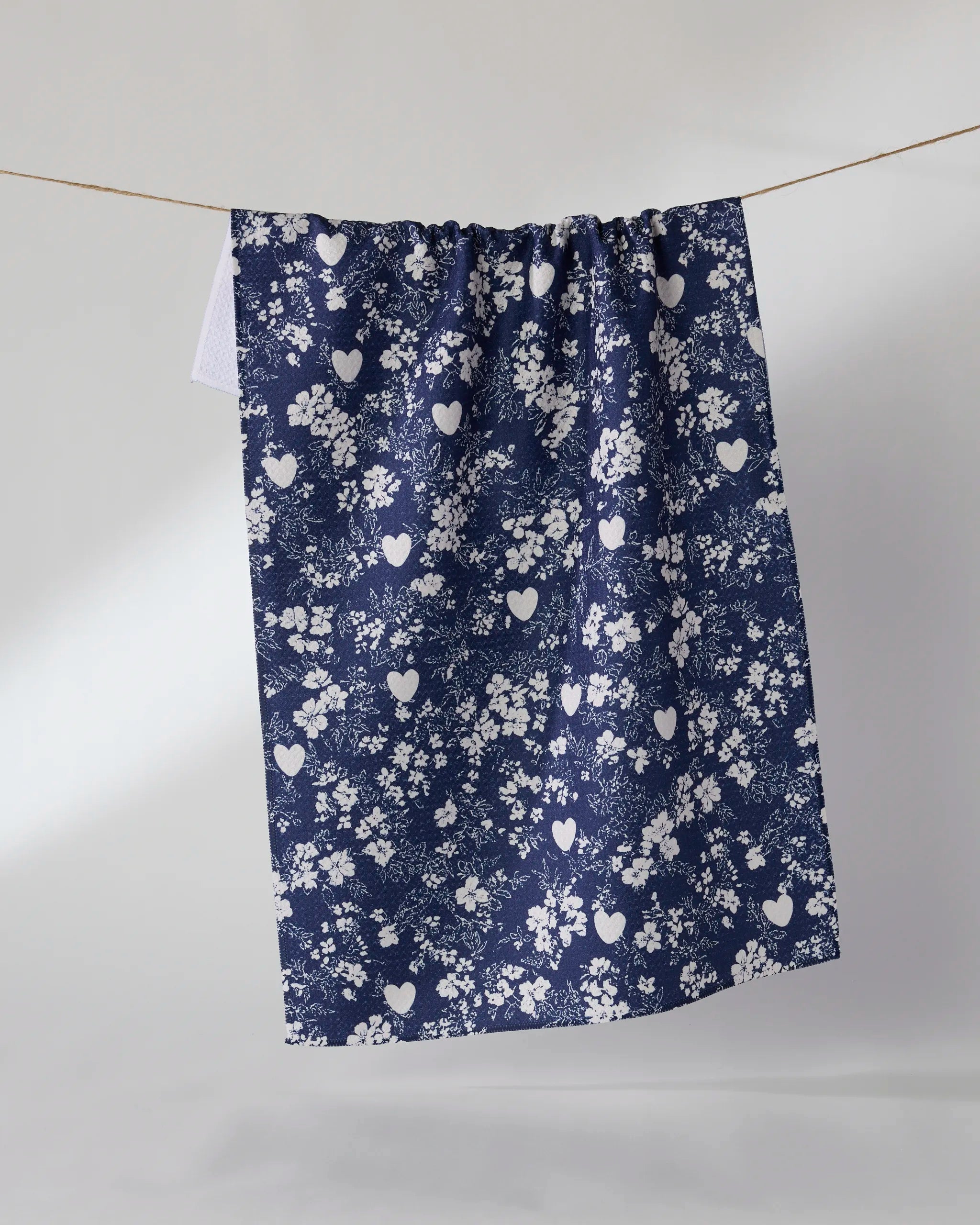 The Geometry "Midnight Posy" tea towel, in navy blue with white heart and floral designs, hangs on a clothesline against a light background.
