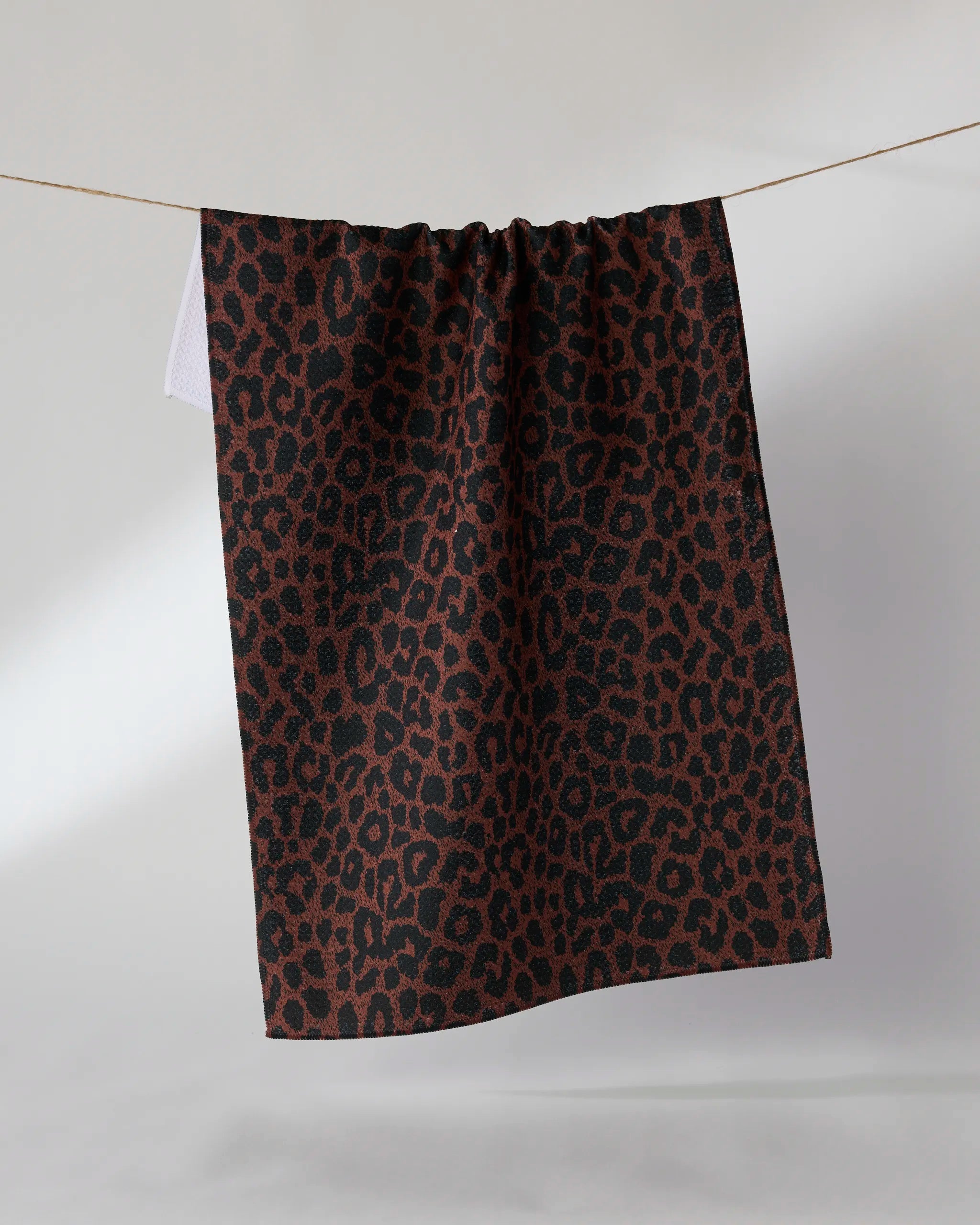 The Geometry Midnight Safari recycled kitchen towel, featuring a brown and black leopard print, hangs over a clothesline against a plain light background.