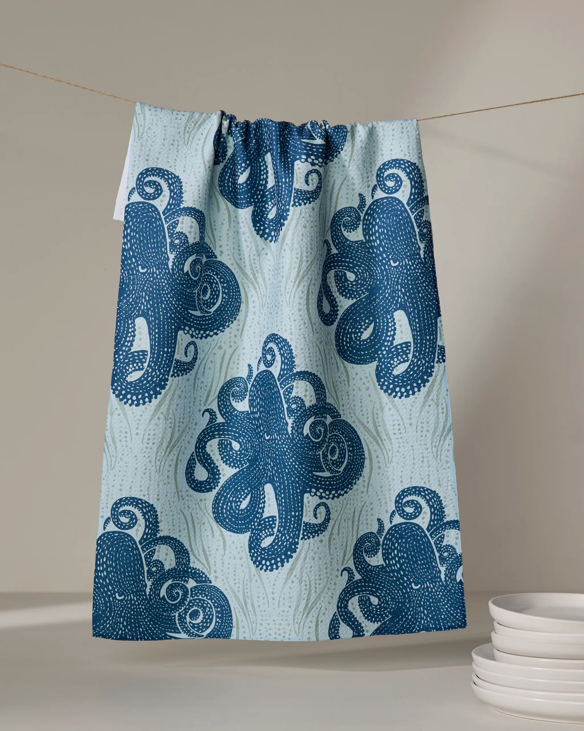 The Katie O'Shea Mighty Octopus tea towel, featuring a blue and white repeating octopus pattern, hangs on a clothesline above a stack of white plates against a softly lit neutral background.