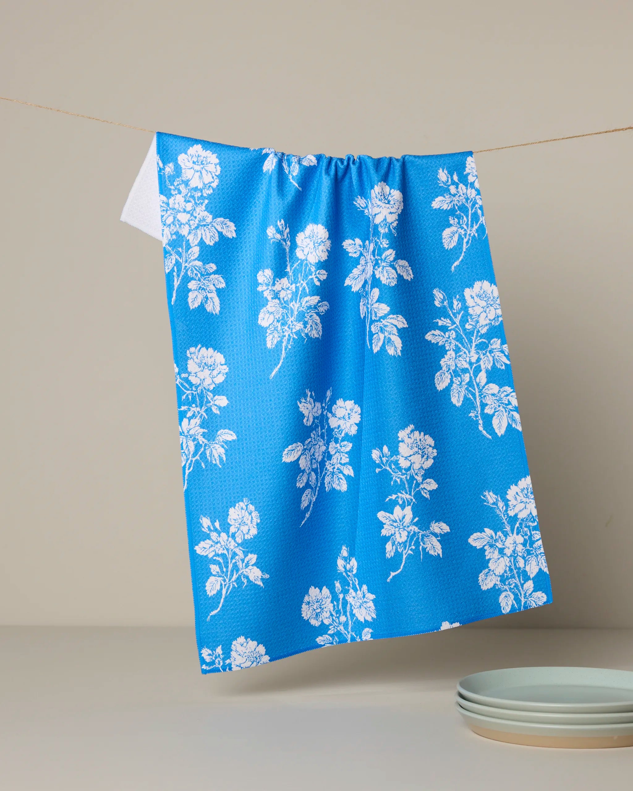The Geometry Milos blue tea towel with a white floral pattern hangs above a beige surface, next to a stack of light blue plates.