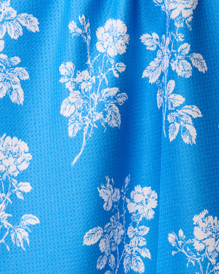 The Milos tea towel by Geometry is a bright blue towel with a textured surface, showcasing an intricate white floral and leaf print scattered across the material.