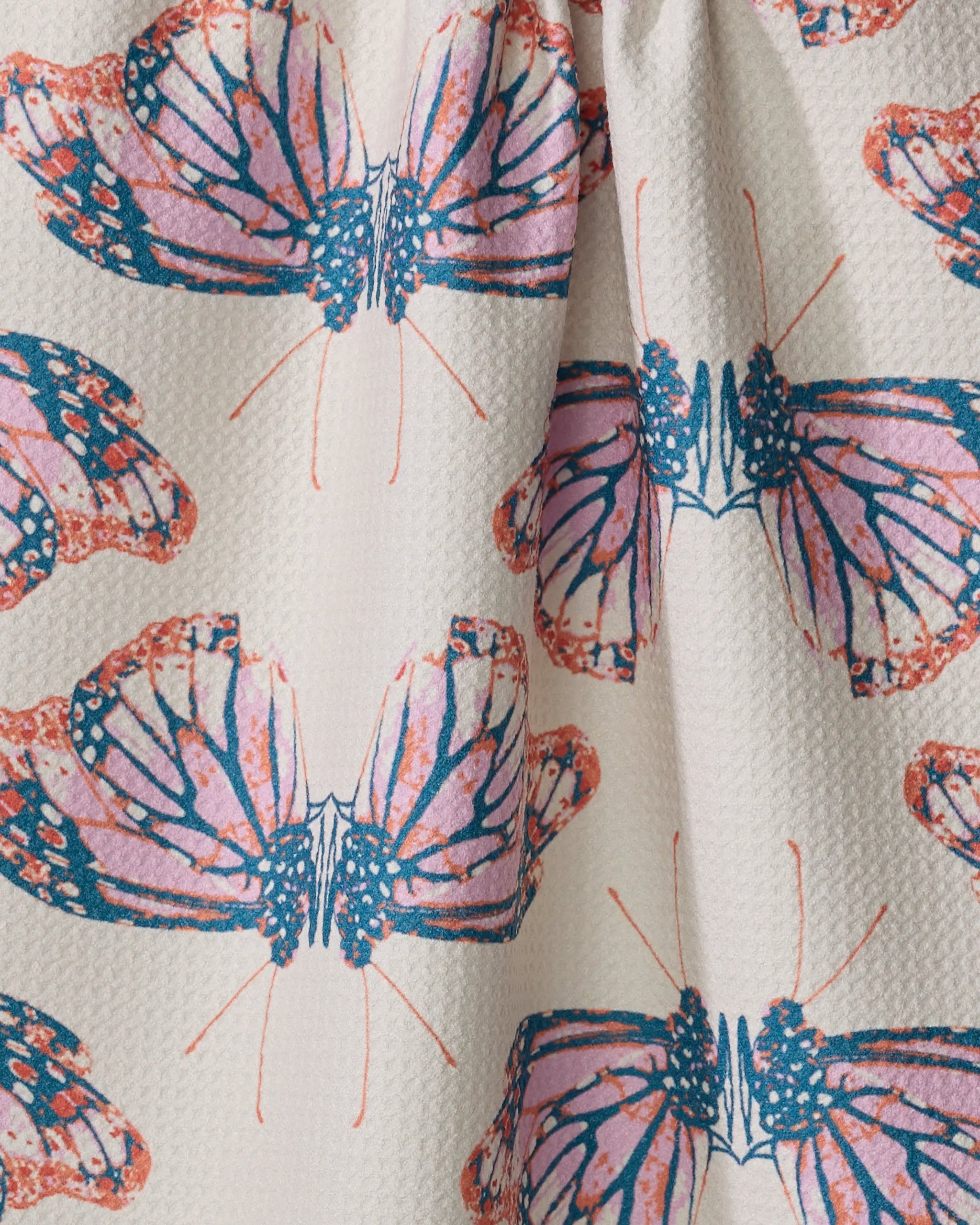 The Mimicry dish towel by Rebecca Woolbright is a white textured, absorbent towel adorned with a repeating pattern of orange, pink, and blue monarch butterflies with open wings.