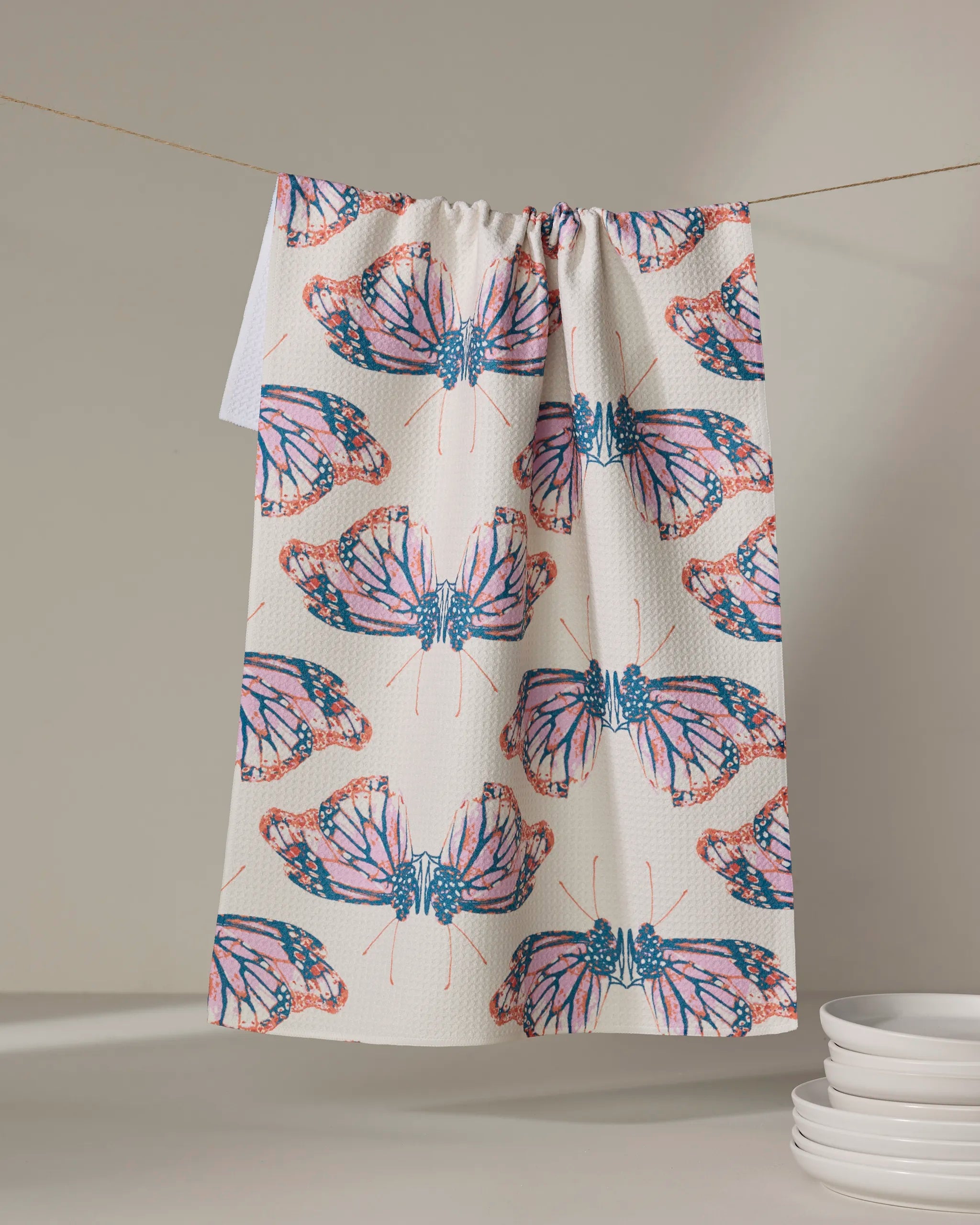 The Mimicry tea towel by Rebecca Woolbright, with vibrant butterfly wings in pink, orange, and blue, hangs on a line above a stack of white plates. The absorbent dish towel adds a pop of color to your kitchen decor.