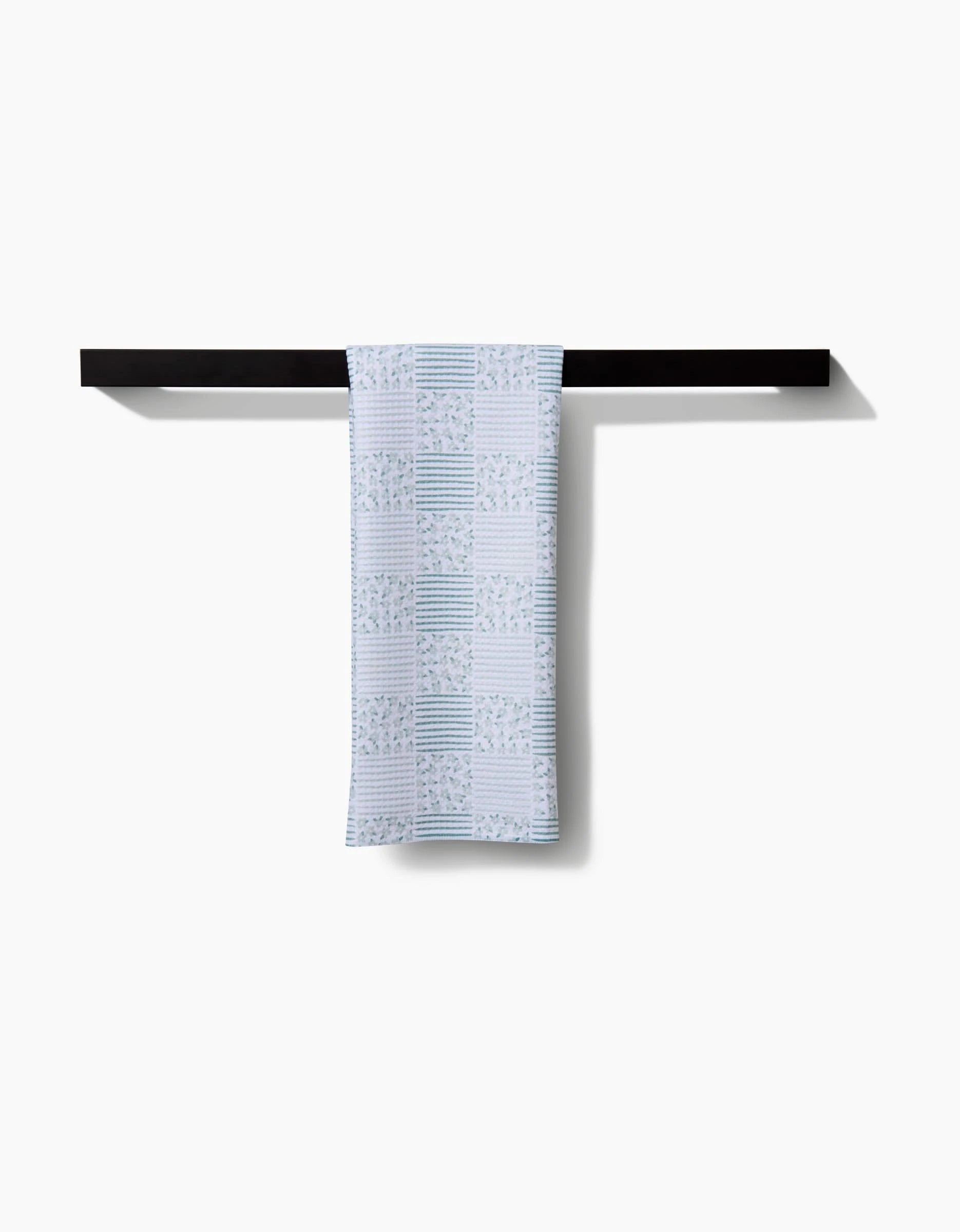 The Hufton Studio Mini Flowers and Stripes Check Green tea towel, made from Geoweave™ fabric, hangs neatly over a minimalist black rod against a plain white background.