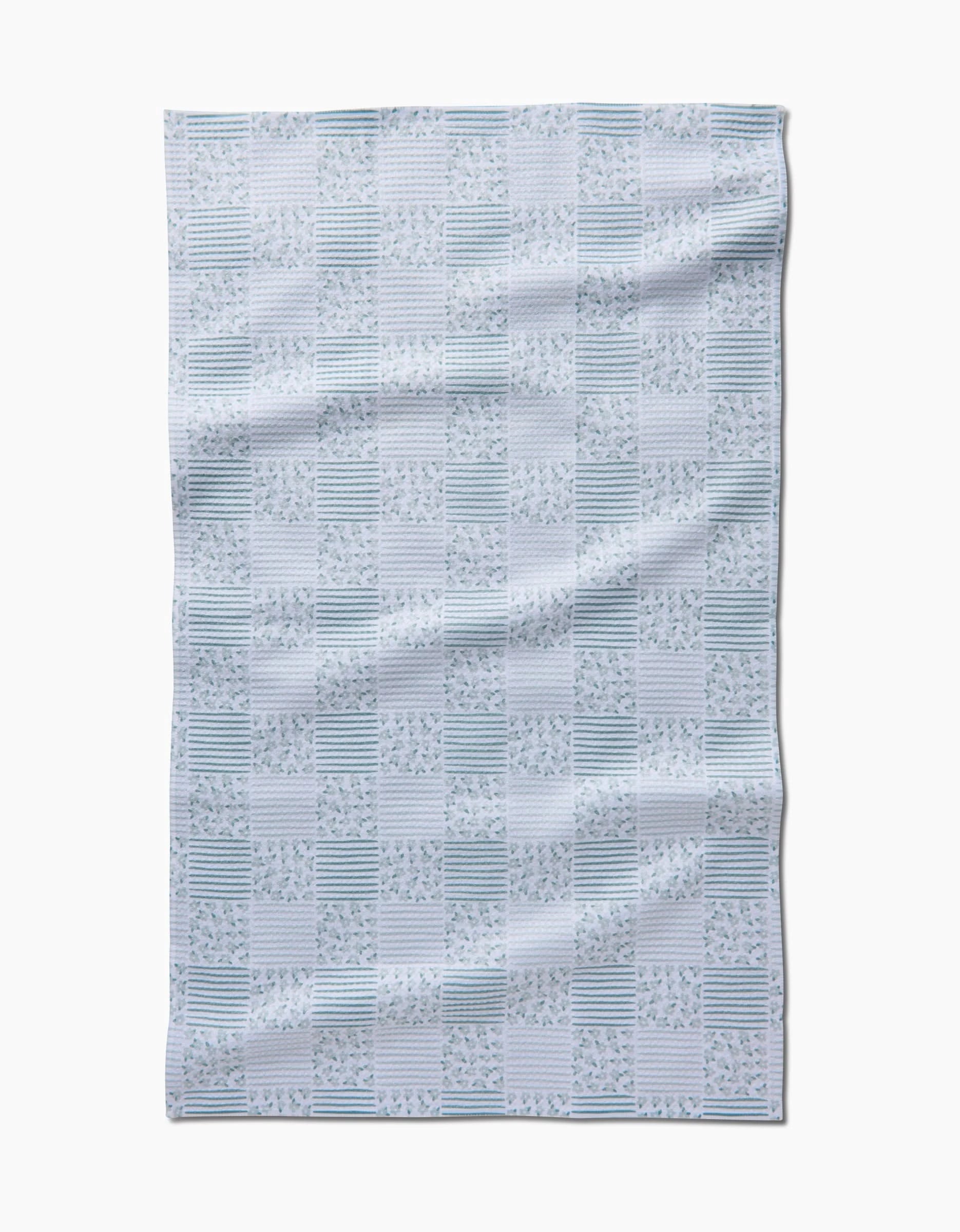 The Mini Flowers and Stripes Check Green tea towel by Hufton Studio features a rectangular white design with green checkered squares, leaf motifs, and stripes. Made from recycled materials for eco-friendly appeal, shown on a white background.