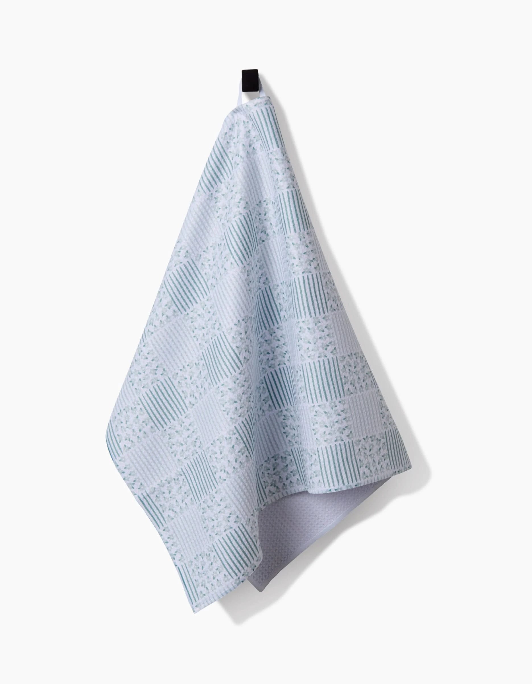The Hufton Studio Mini Flowers and Stripes Check Green tea towel, featuring checkered, striped, and floral patterns in green hues on Geoweave™ fabric, hangs from a black hook against a white background.