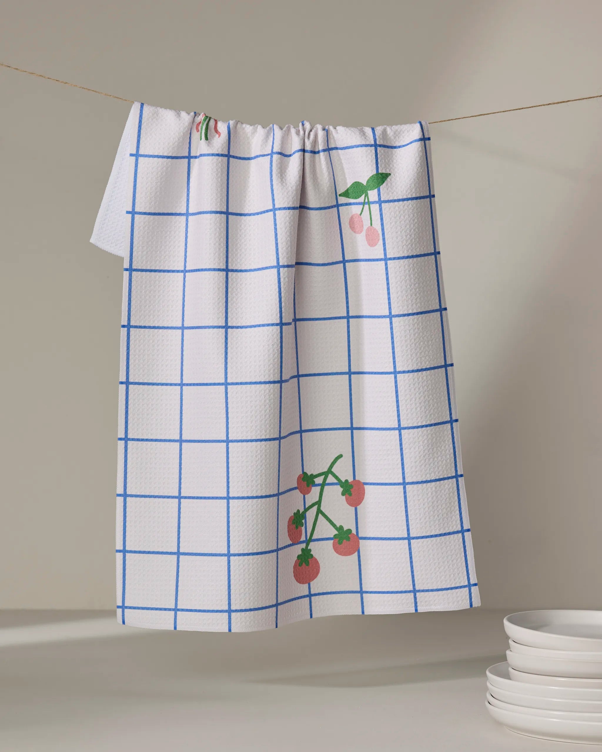 The Mini Harvest dish towel by Letters By Shells, featuring a blue grid and cherry print, hangs on a clothesline above white plates, adding charm and function to your kitchen.