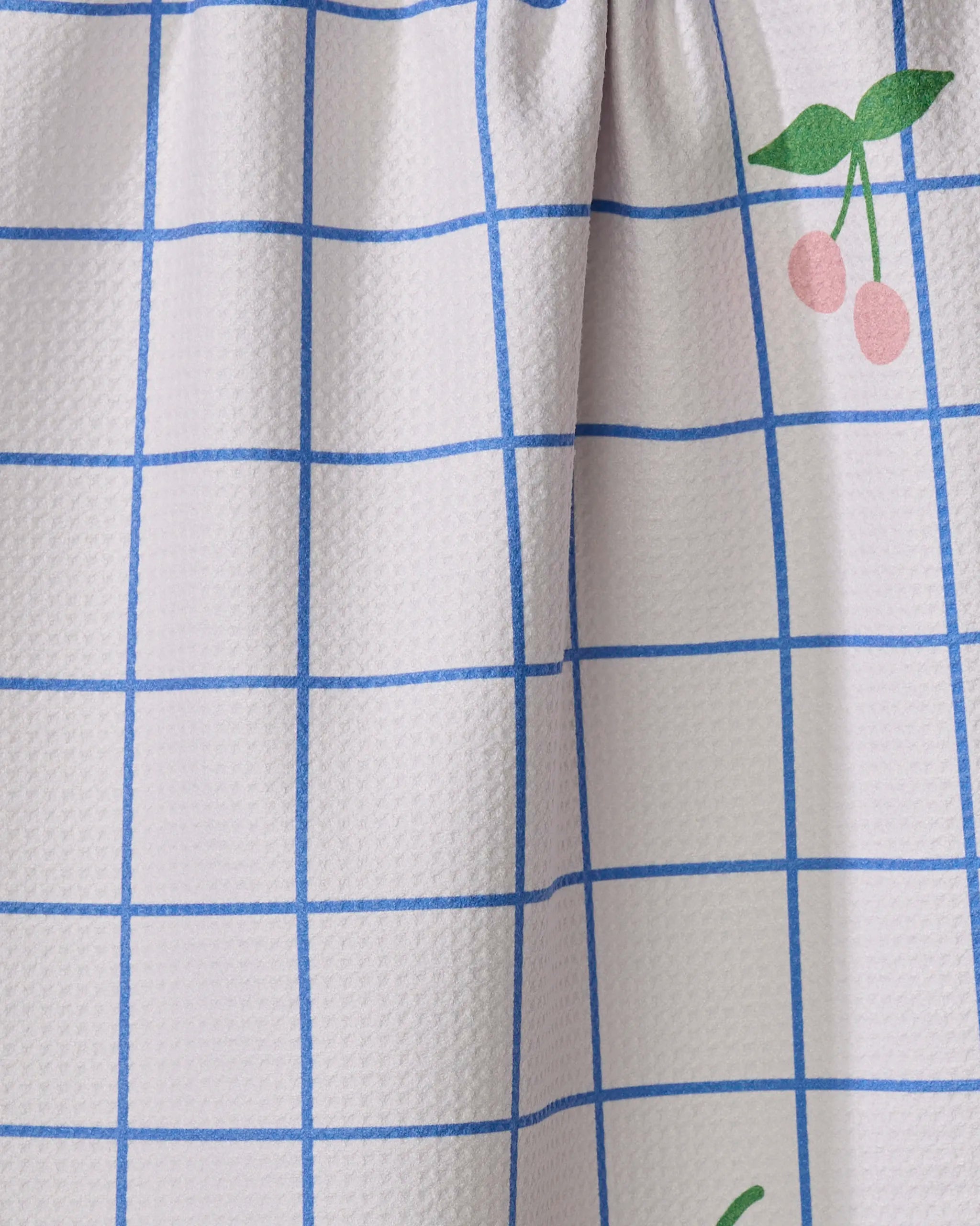 The Mini Harvest tea towel by Letters By Shells features white textured fabric, a blue grid pattern, and a playful small illustration of two pink cherries with green leaves in the upper right corner.