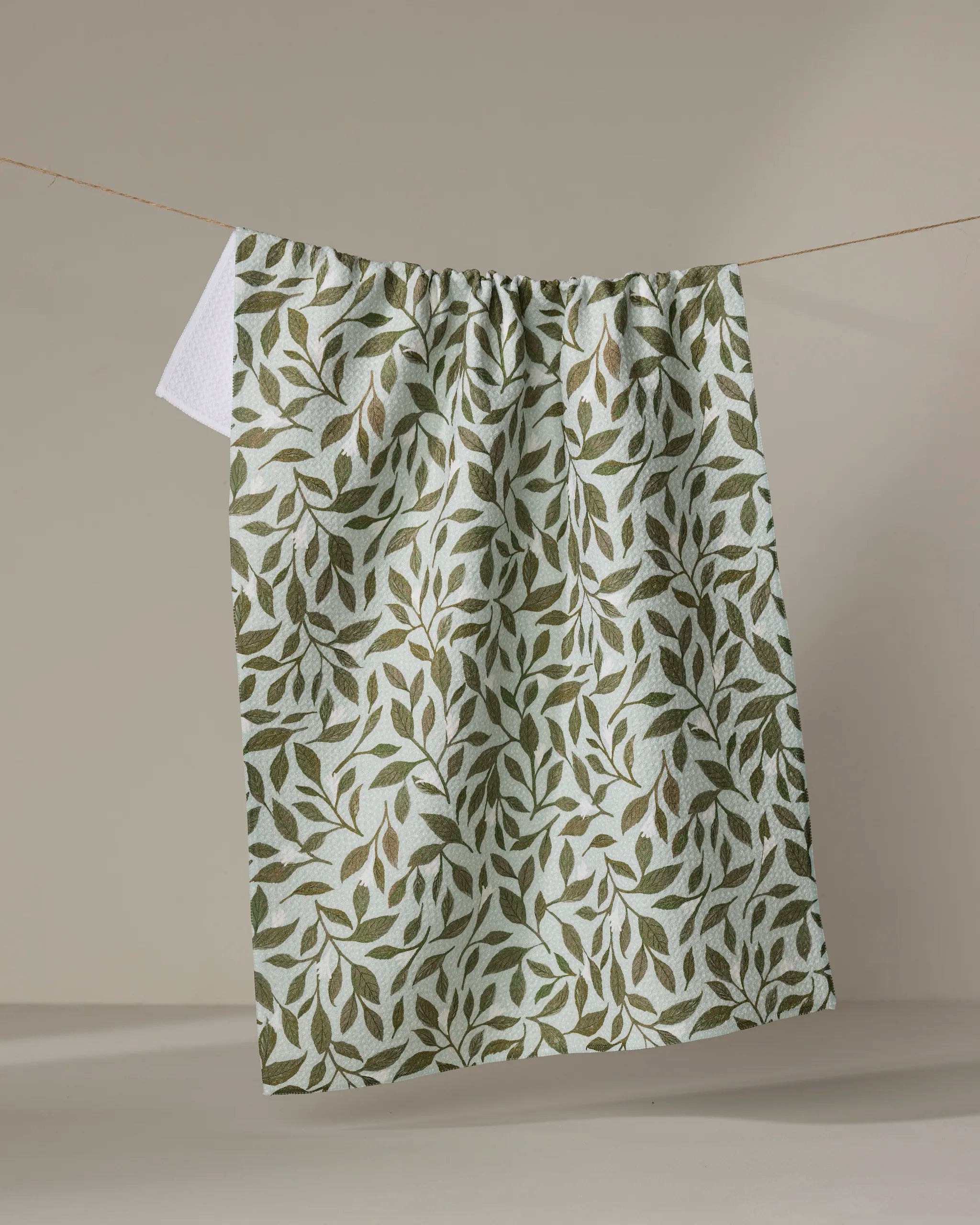 The Katie O'Shea "Mint Leaves" kitchen towel, featuring a quick-drying, eco-friendly design and green leafy pattern, hangs on a clothesline against a neutral beige background.