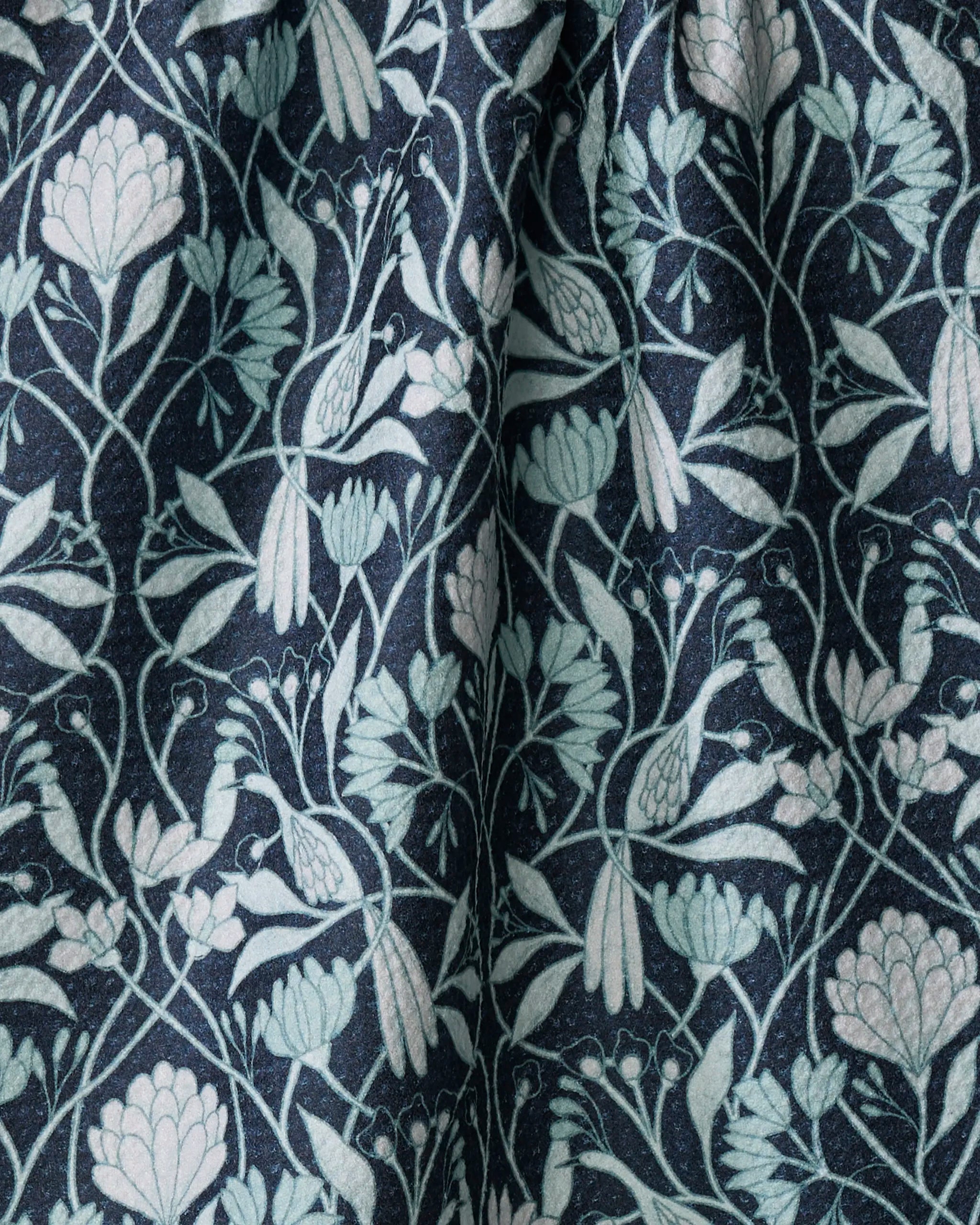 The Mirielle kitchen towel by Amy MacCready features dark fabric with a symmetrical design of light blue and pale green flowers, leaves, and birds. Made from recycled materials, it has a slightly wrinkled, textured botanical surface.