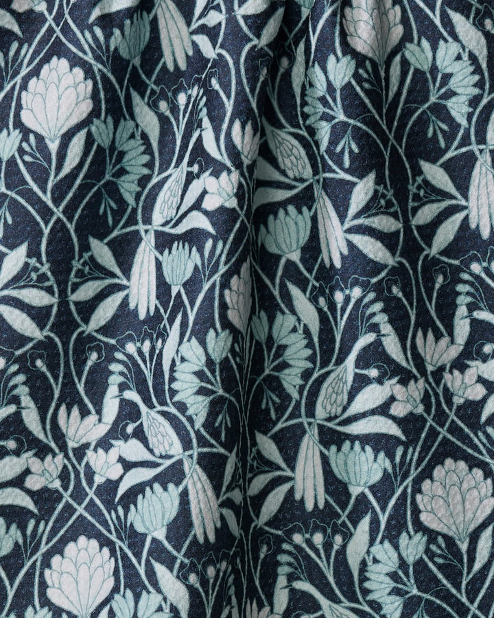 The Mirielle kitchen towel by Amy MacCready features dark fabric with a symmetrical design of light blue and pale green flowers, leaves, and birds. Made from recycled materials, it has a slightly wrinkled, textured botanical surface.