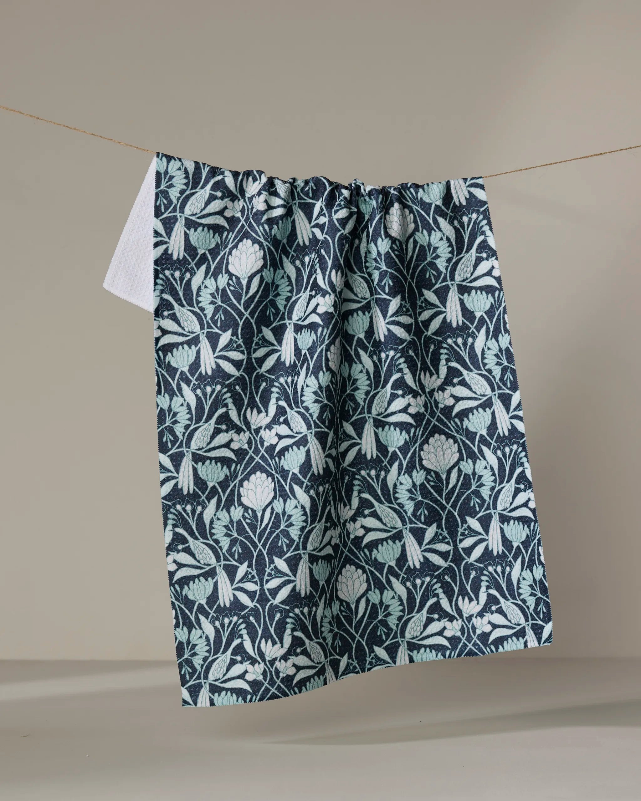 The Mirielle towel by Amy MacCready, featuring a dark blue quick-dry fabric with a light blue floral pattern, hangs from a clothesline against a neutral background.