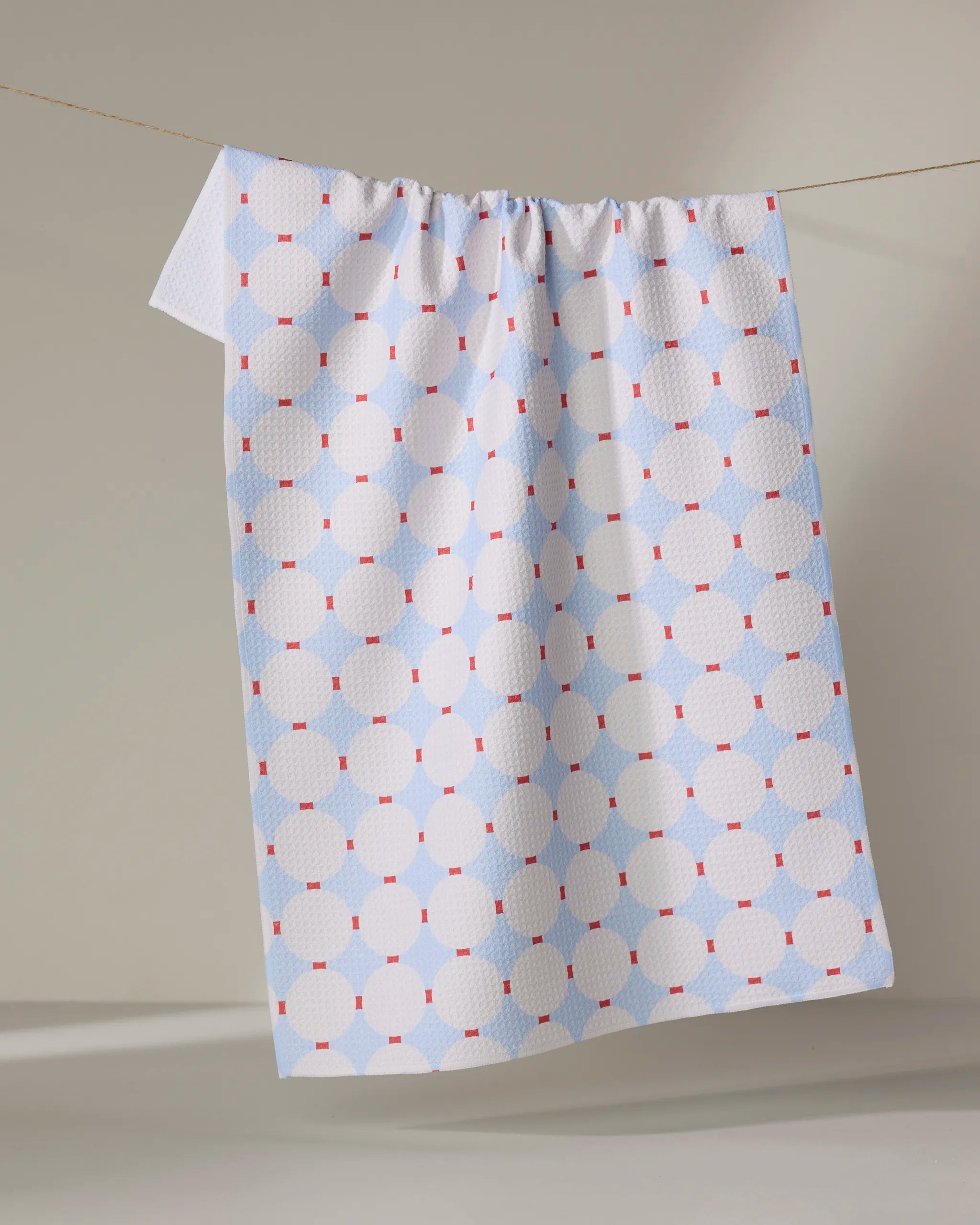 The Happy Hemera Mist Blue Oasis tea towel, featuring a geometric pattern in white and light blue with red squares, made from recycled materials, hangs on a clothesline against a neutral background.