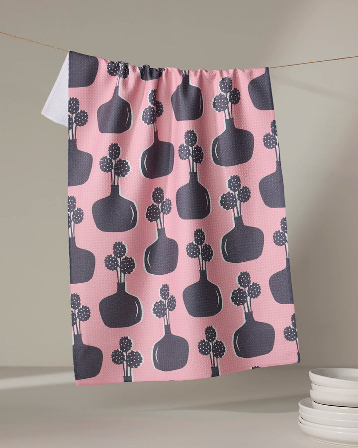The "Modern Bouquet" tea towel by Lizzie Clark features a pink quick-drying design with dark vases and floral motifs. It hangs on a line with a folded white corner, while white plates are stacked nearby.