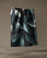 The Geometry Modern Organic tea towel, quick-drying and dark-colored with a large blue-green leaf pattern, hangs on a string against a plain background.