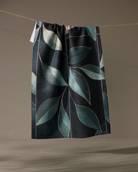 The Geometry Modern Organic tea towel, quick-drying and dark-colored with a large blue-green leaf pattern, hangs on a string against a plain background.