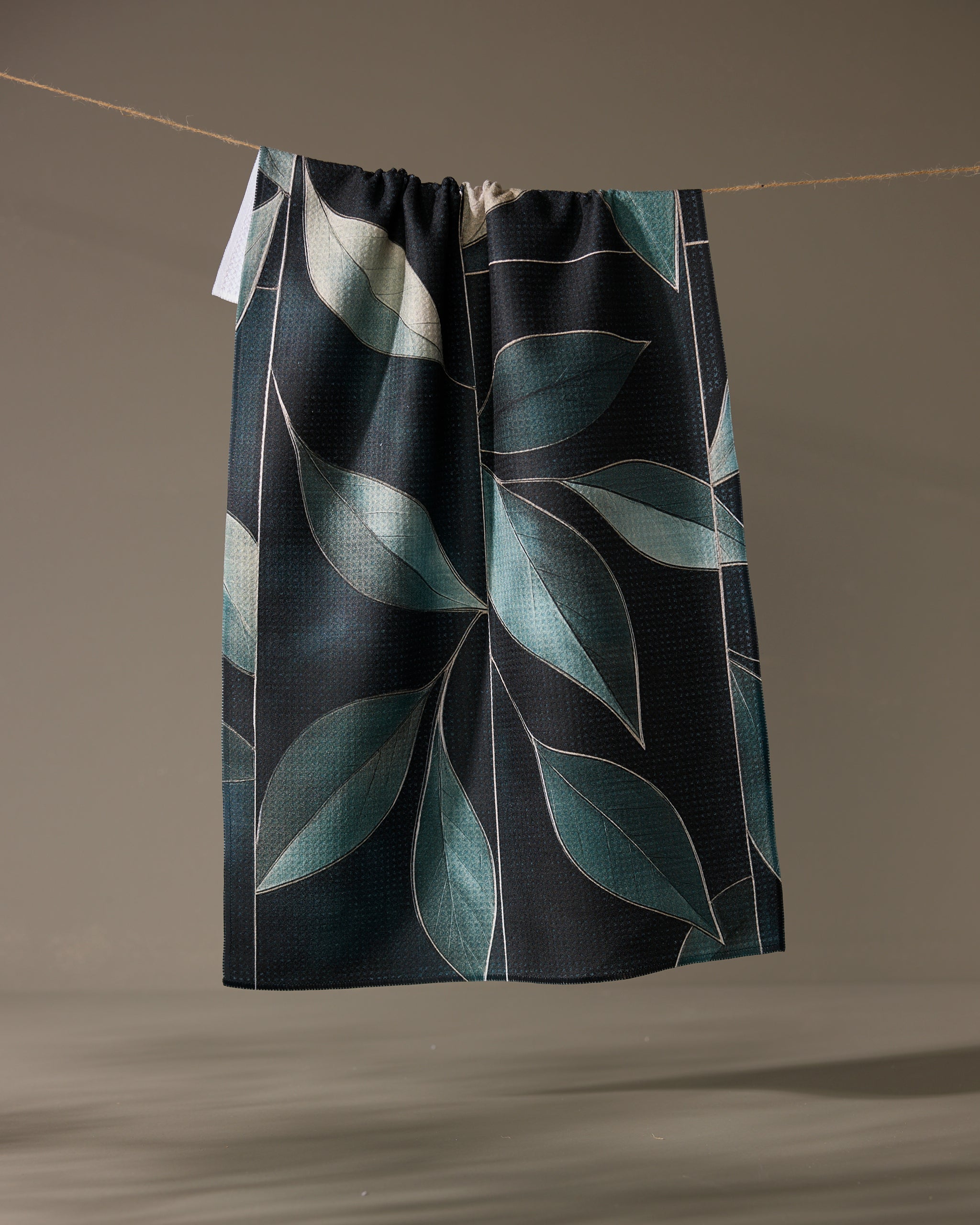 The Geometry Modern Organic tea towel, quick-drying and dark-colored with a large blue-green leaf pattern, hangs on a string against a plain background.