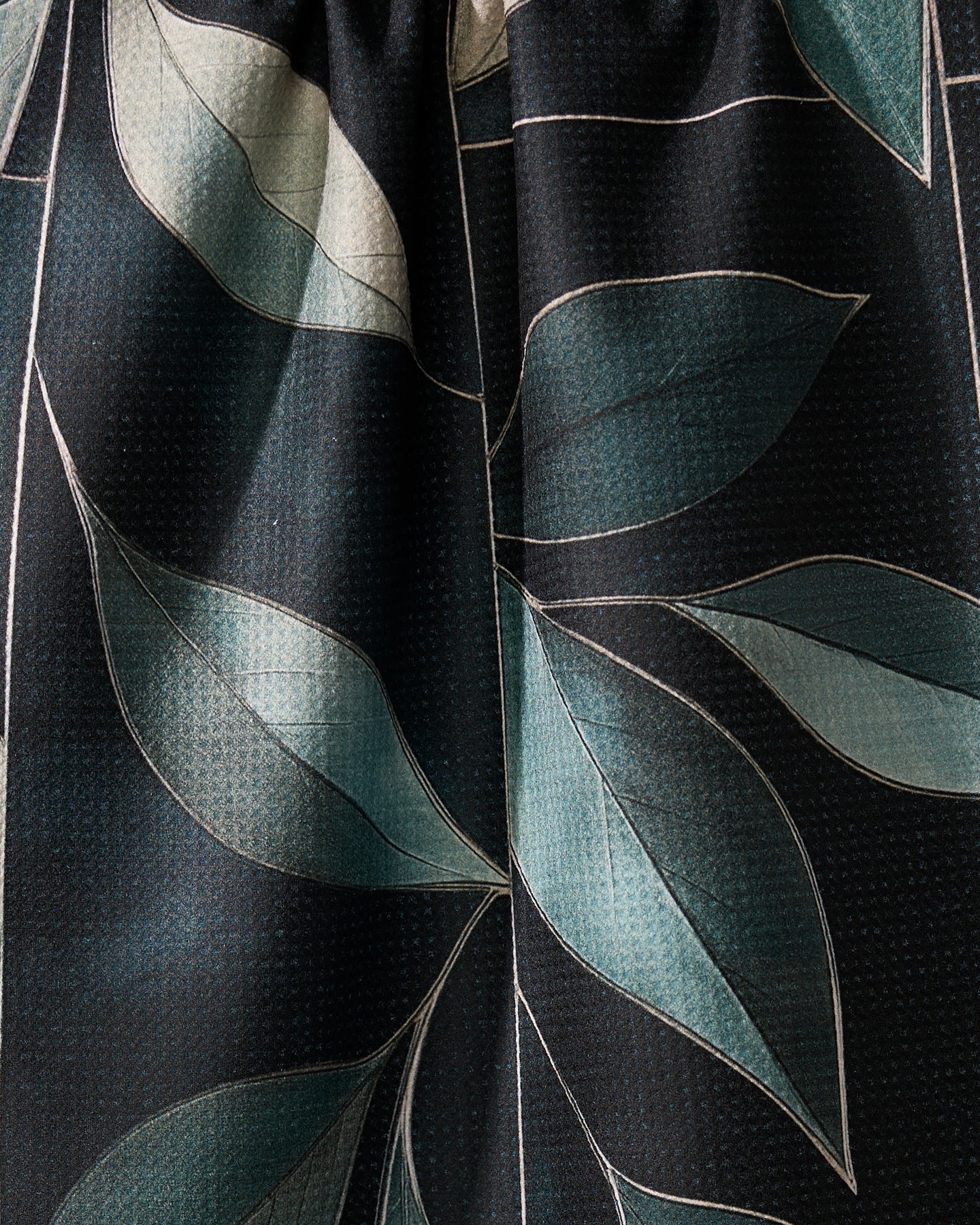 A close-up of Modern Organic by Geometry shows dark fabric with large teal and light green leaves outlined in white. This recycled kitchen towel has a subtle textured surface, combining bold, modern botanical design with sustainability.
