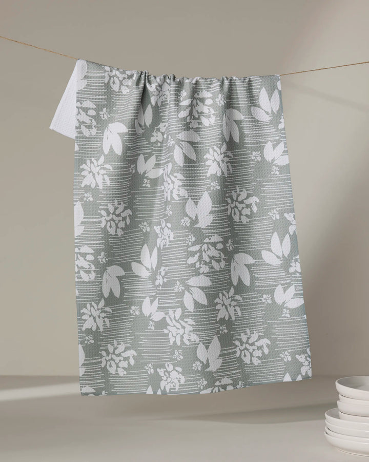 The Toboggan Avenue "Moments" kitchen tea towel, featuring a gray and white floral and leaf pattern made from recycled materials, hangs on a clothesline near a stack of white plates against a neutral background.