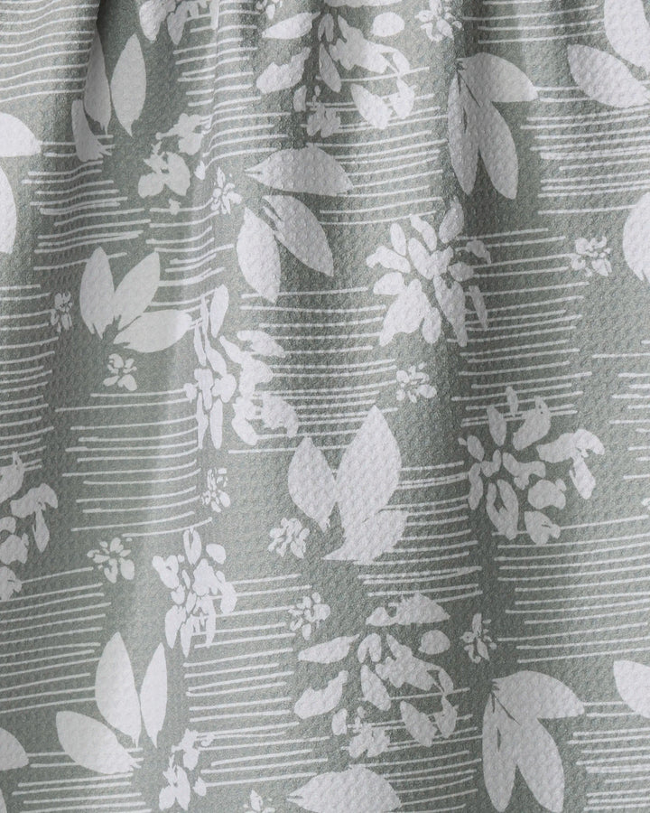 Close-up of the Moments kitchen tea towel by Toboggan Avenue, crafted from recycled materials with a white leaf and flower pattern on light gray, featuring horizontal lines and gentle folds.