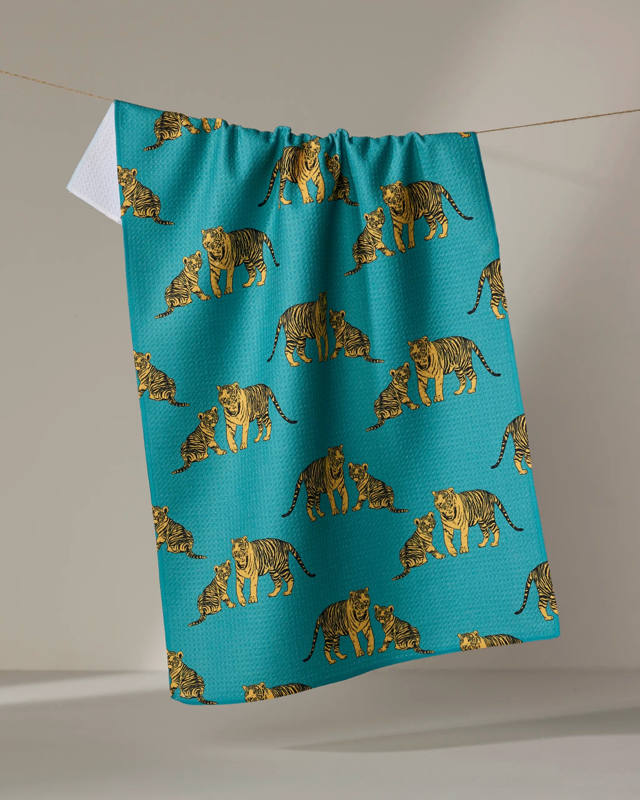 The "Mommy and Me" tea towel by Sarah Holden is a teal ultra-absorbent towel with illustrated tigers and cubs in a repeating pattern, shown hanging on a clothesline against a neutral background.