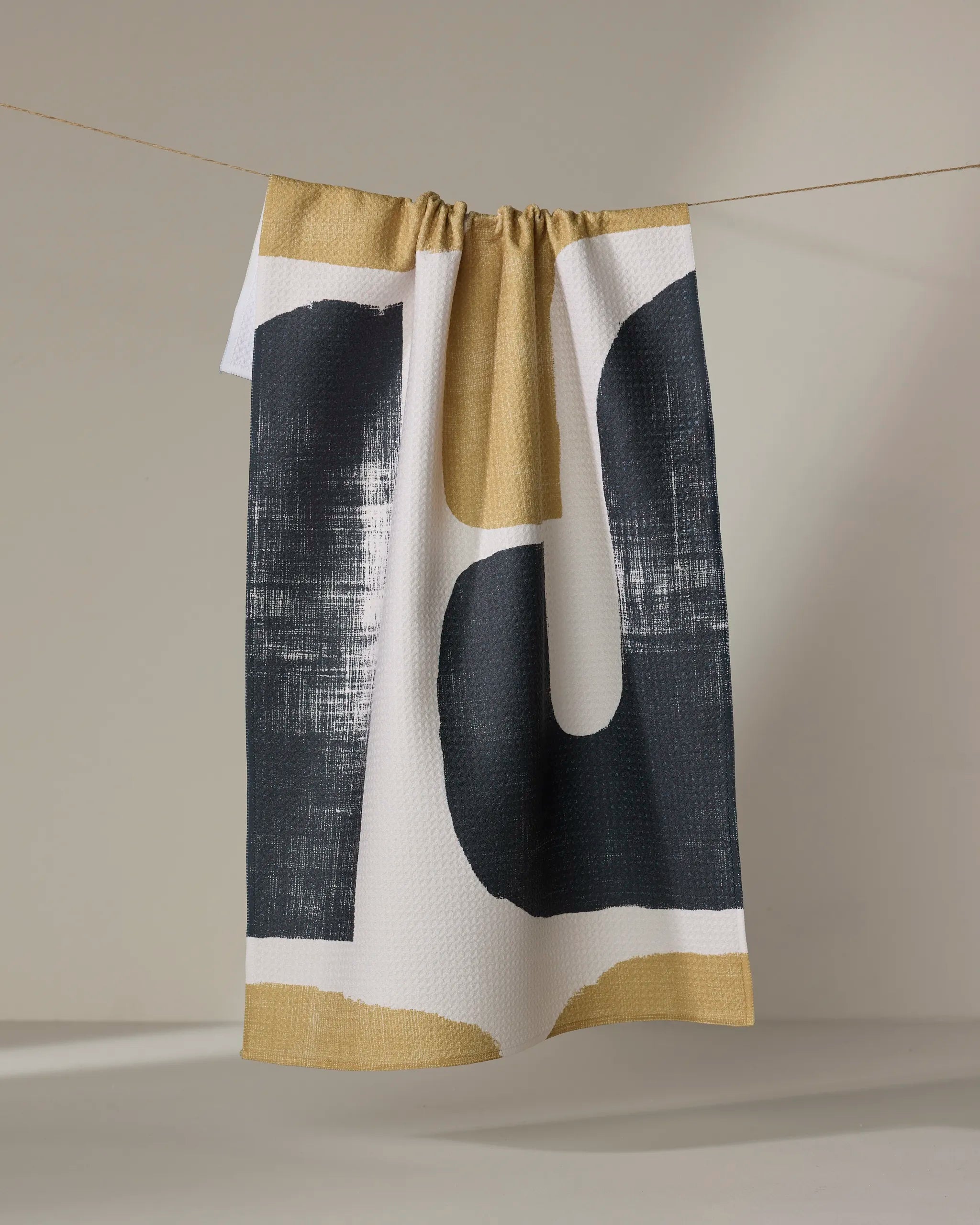 The Geometry Mono towel, featuring an abstract black and gold geometric design, hangs from a string against a neutral background.
