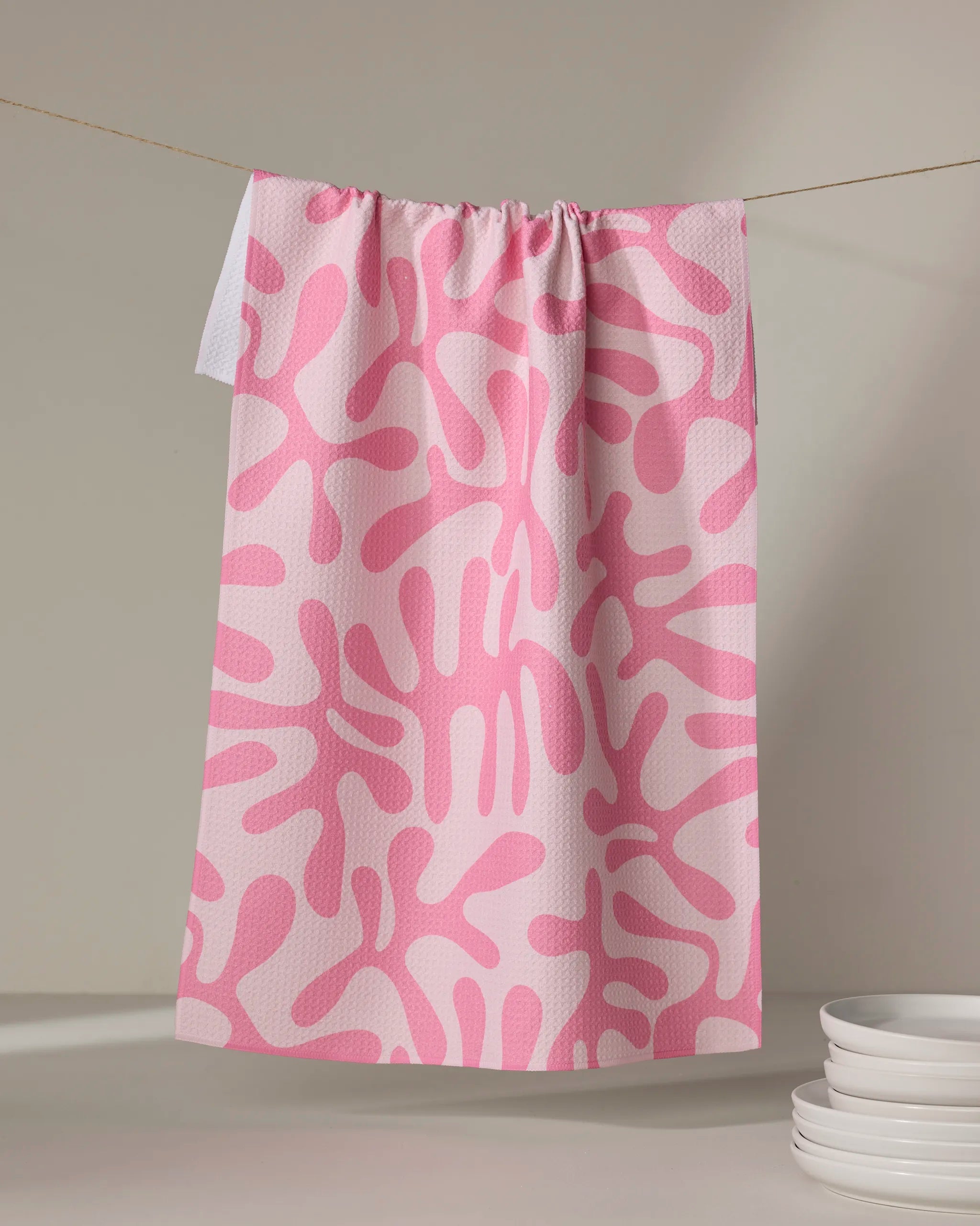 The "Monochrome Morning" tea towel by Marina Solodka, featuring an abstract pink and white splatter pattern, hangs on a clothesline. Made from recycled materials, it dries quickly. White plates are stacked in the lower right corner on a neutral background.