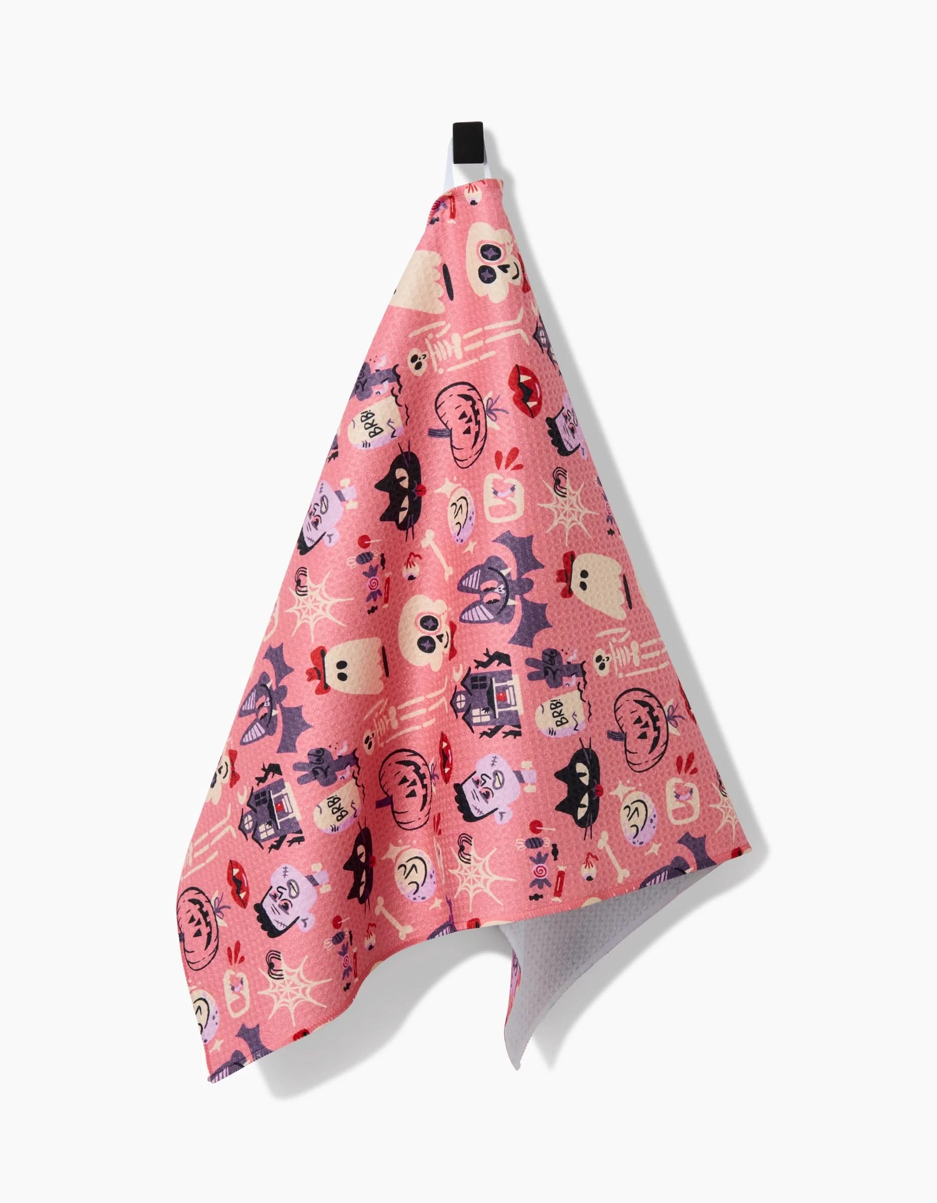 The Carly Berry Monstermash kitchen towel is pink, made from recycled materials, and features playful cartoon faces, animals, and doodles. It hangs by a black loop against a white background.