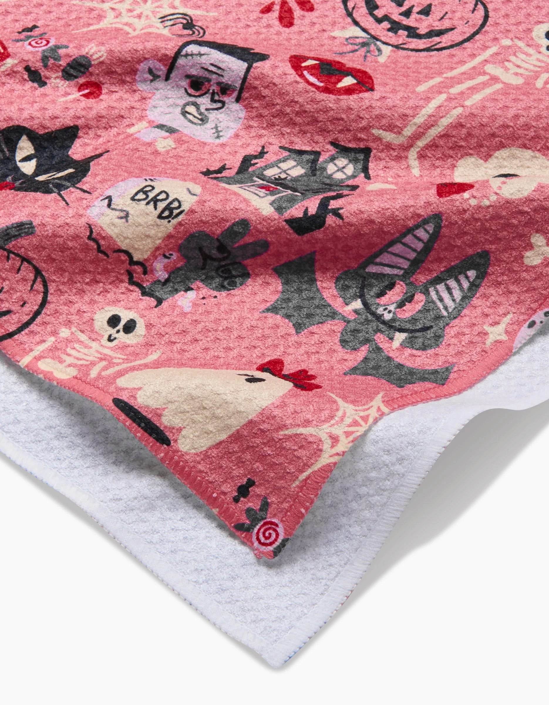 A close-up of Carly Berrys Monstermash pink kitchen towel, made from recycled materials and decorated with playful Halloween icons—bats, pumpkins, tombstones, and skulls—with a white underside peeking through.