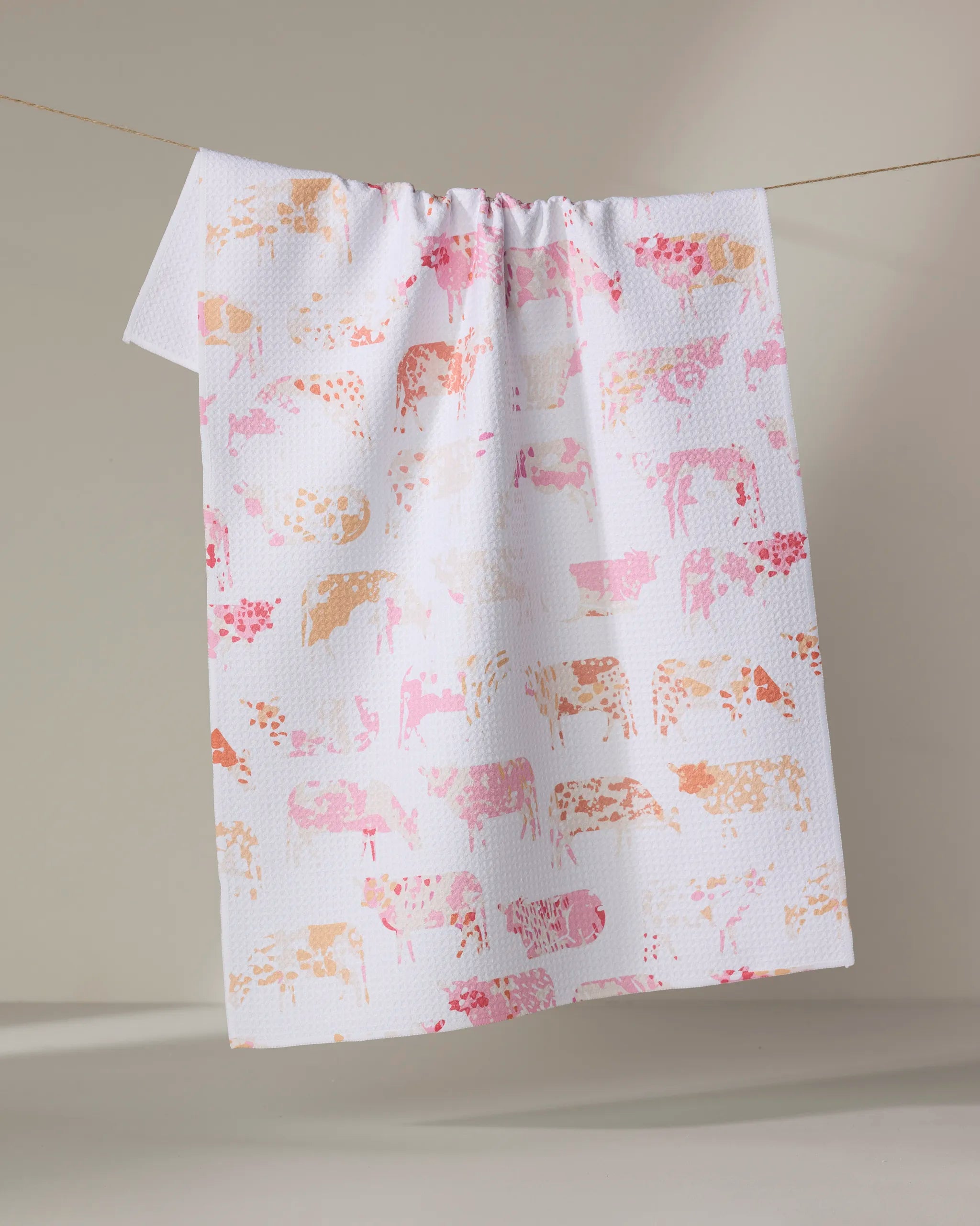The "Moods" tea towel by Elisabeth Wing features a pink and orange cow pattern on white, made from recycled materials, and is shown draped over a clothesline against a neutral background.
