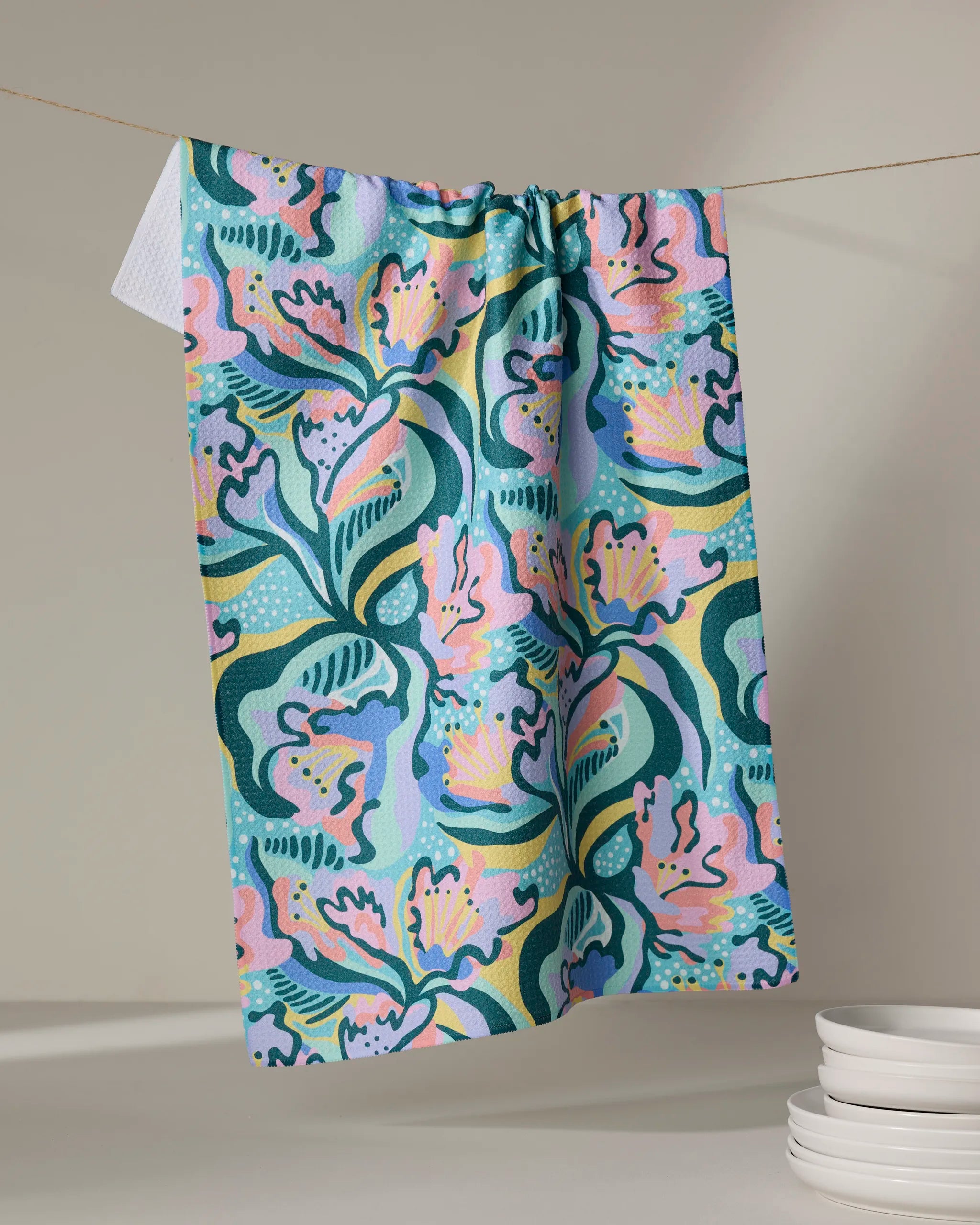 The Moody Flowers recycled kitchen towel by Masha Volnova, featuring an abstract floral pattern in pink, blue, green, and yellow, hangs on a line beside stacked white dishes on a neutral surface.