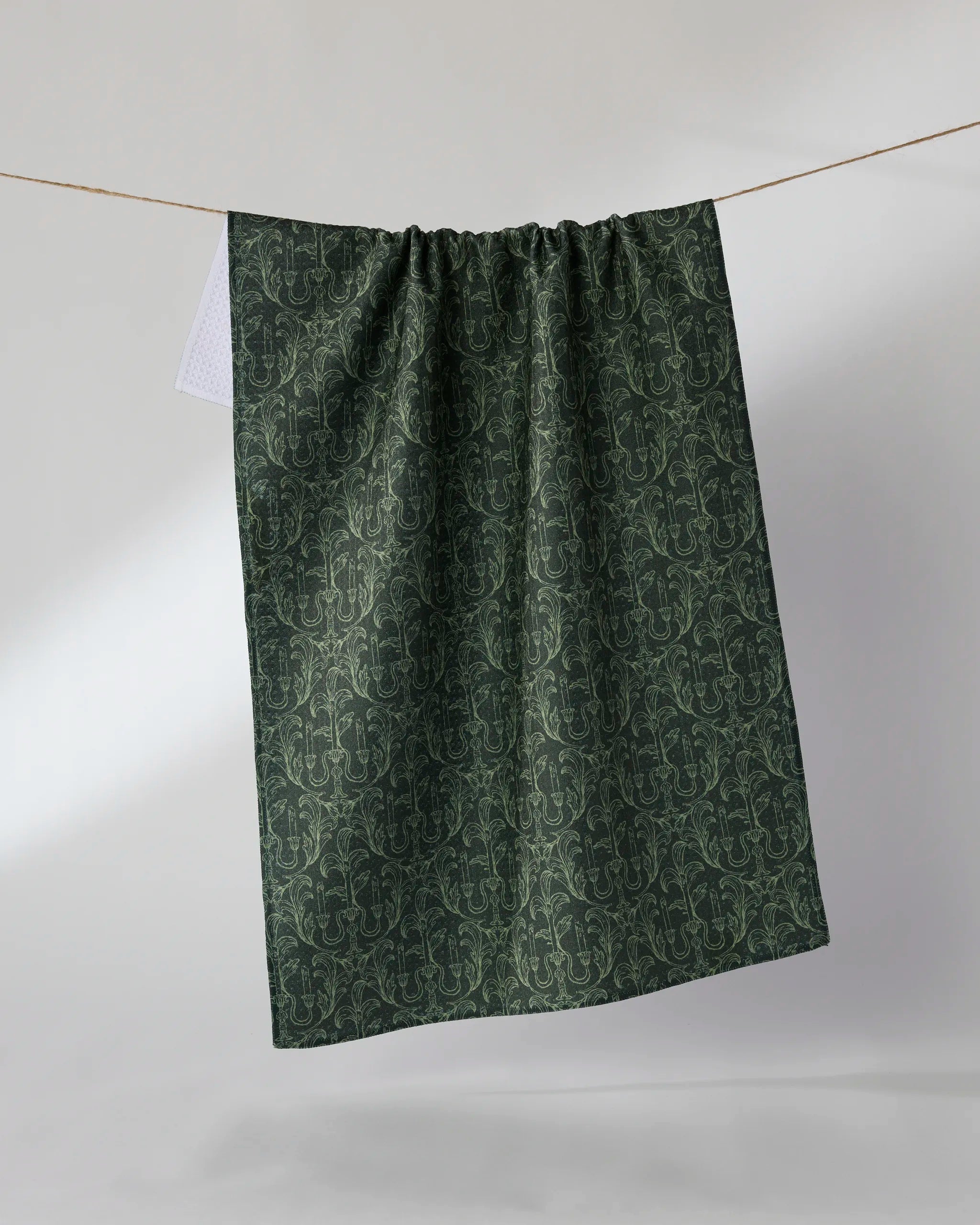 The Geometry "Moody Manor" recycled kitchen towel in dark green, featuring an intricate floral pattern, hangs on a clothesline against a light, neutral background.