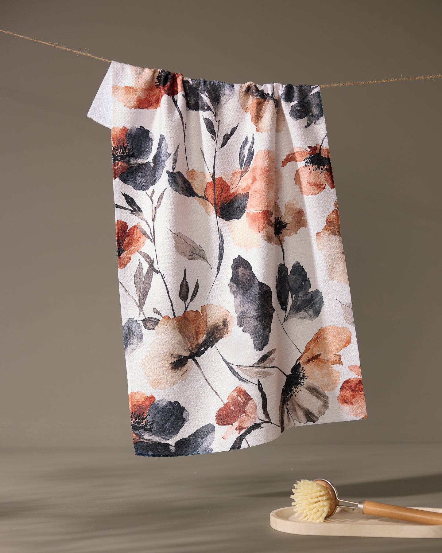 The Moody Poppy tea towel by Geometry, featuring orange, black, and gray floral designs on recycled fabric, hangs on a string above a wooden dish brush set on a small round surface against a plain neutral background.