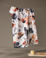 The Moody Poppy tea towel by Geometry, featuring orange, black, and gray floral designs on recycled fabric, hangs on a string above a wooden dish brush set on a small round surface against a plain neutral background.