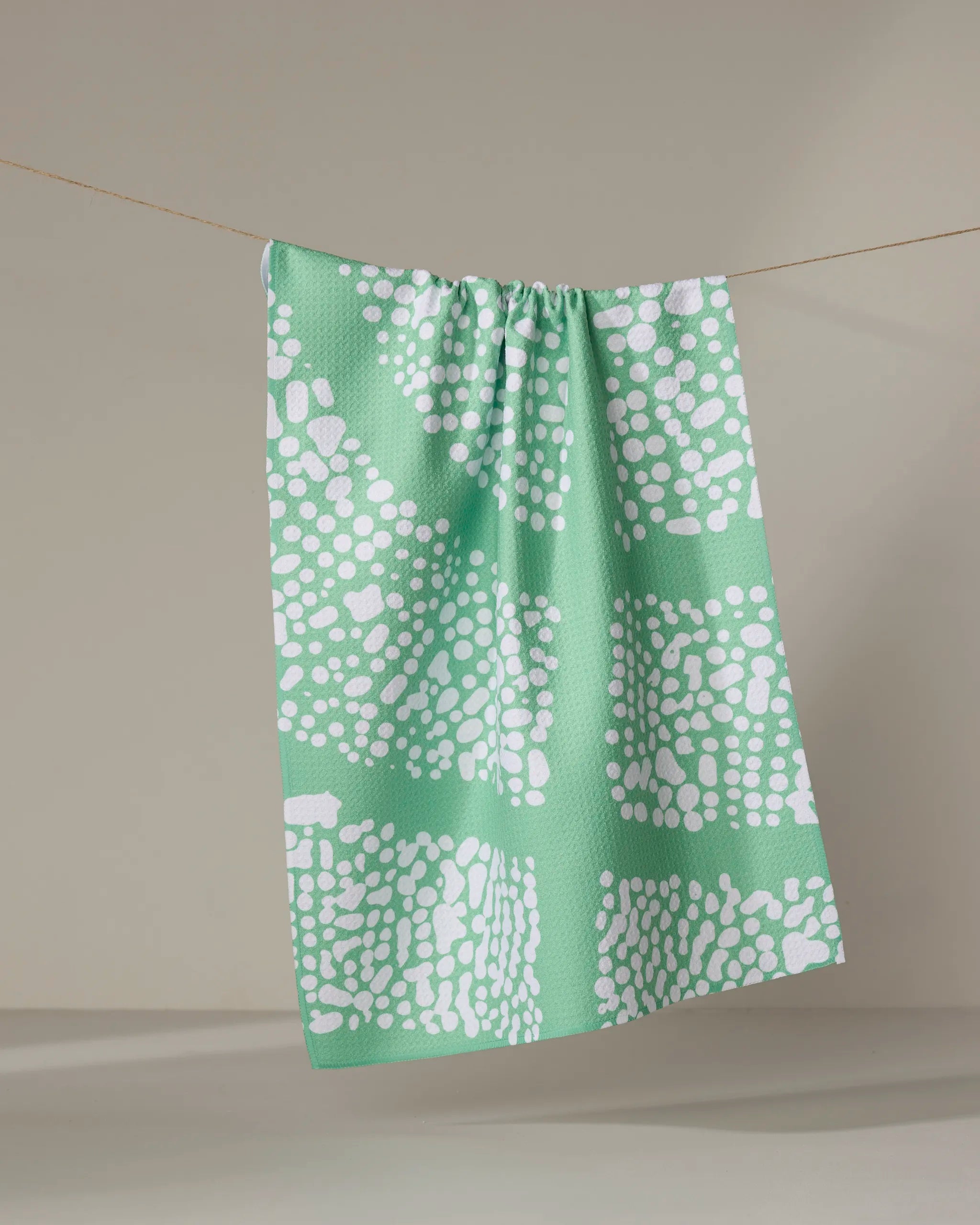 The Sissy Moon "Moon Rock" dish towel in light green features a white dotted abstract pattern and hangs on a thin string against a plain, neutral background.