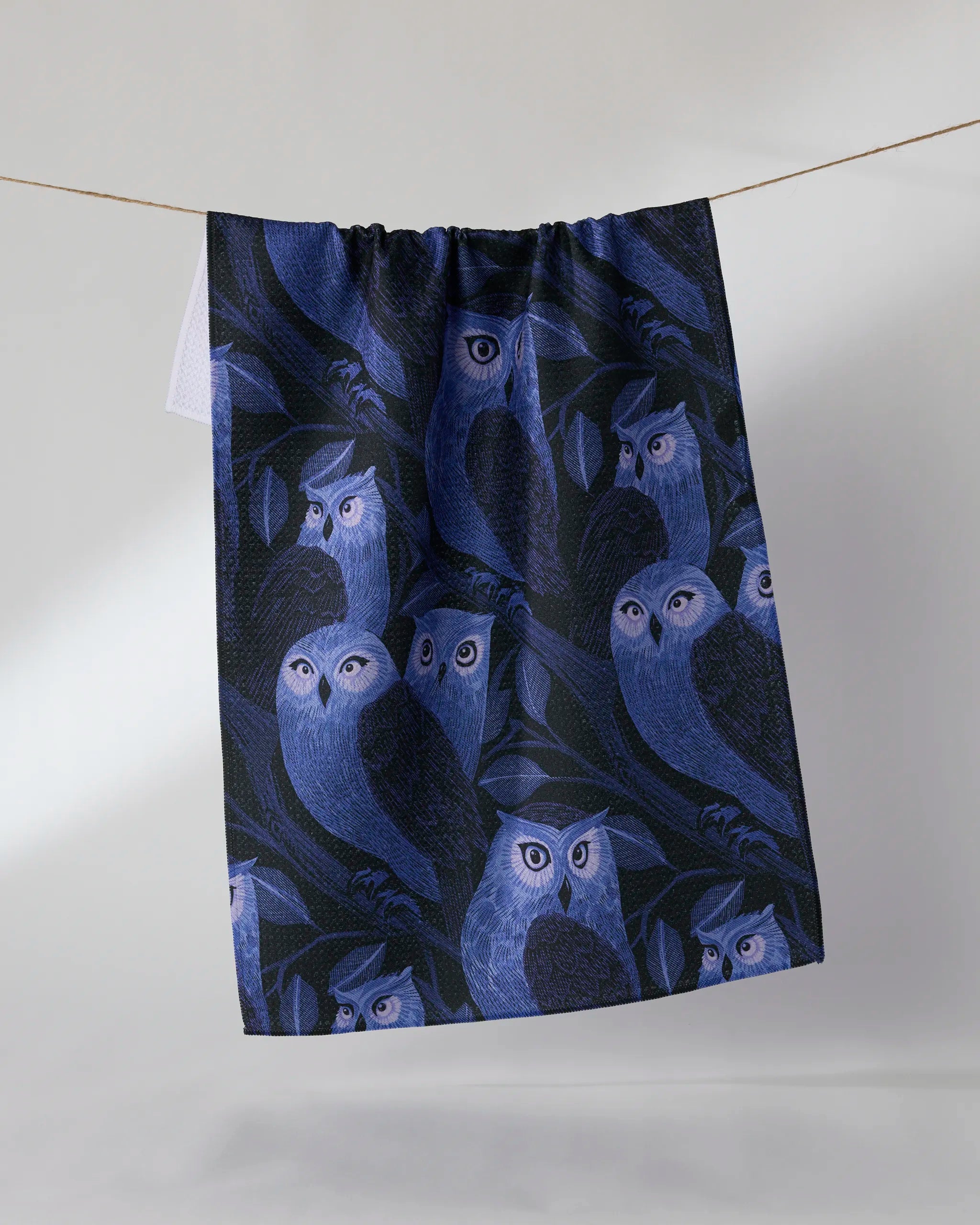 The Geometry Moonfeather tea towel, featuring blue and purple owls among dark blue leaves, hangs from a string against a plain light background.