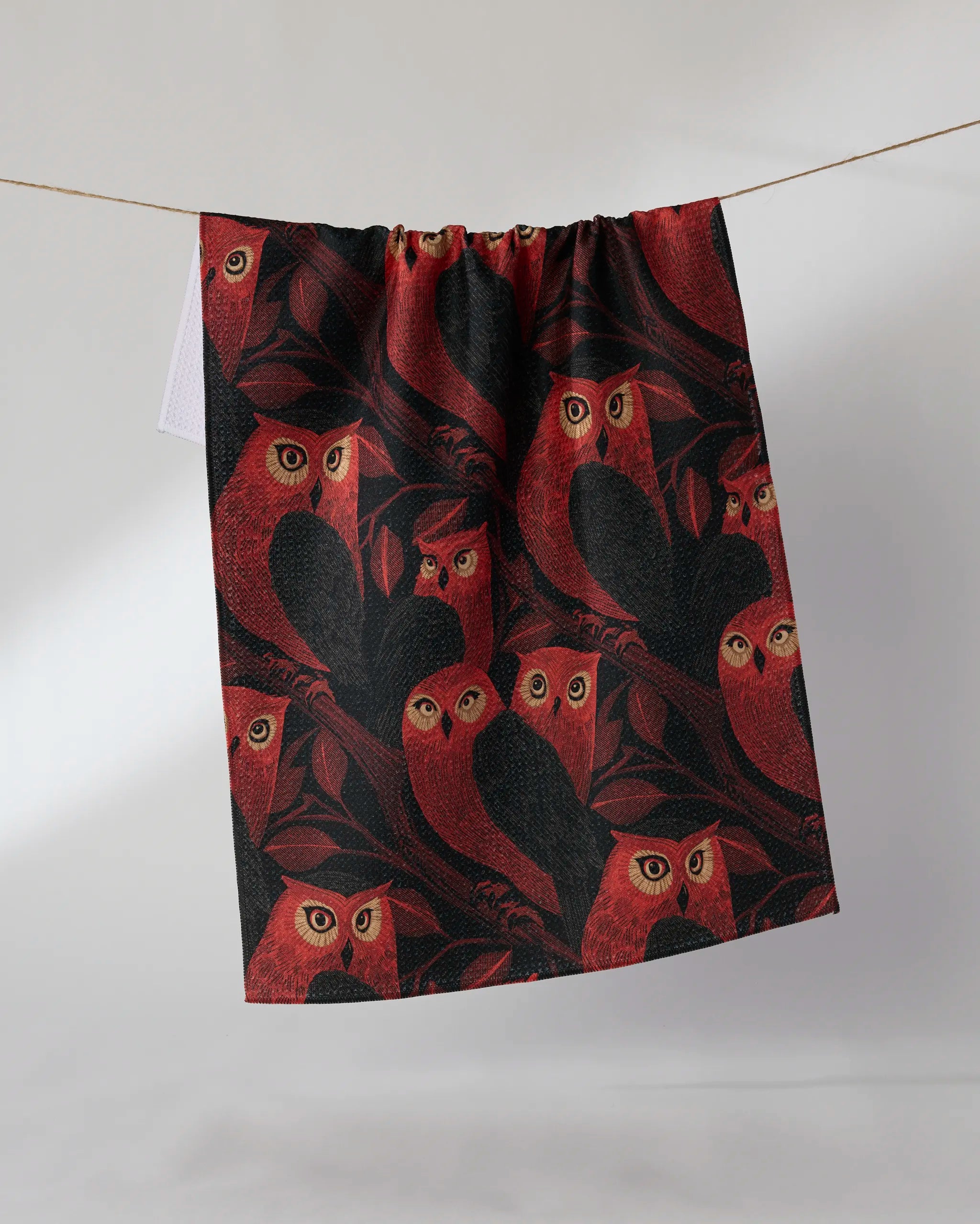 The Geometry Moonfeather kitchen towel, featuring a red and black owl print on branches, hangs from a clothesline against a simple light background.