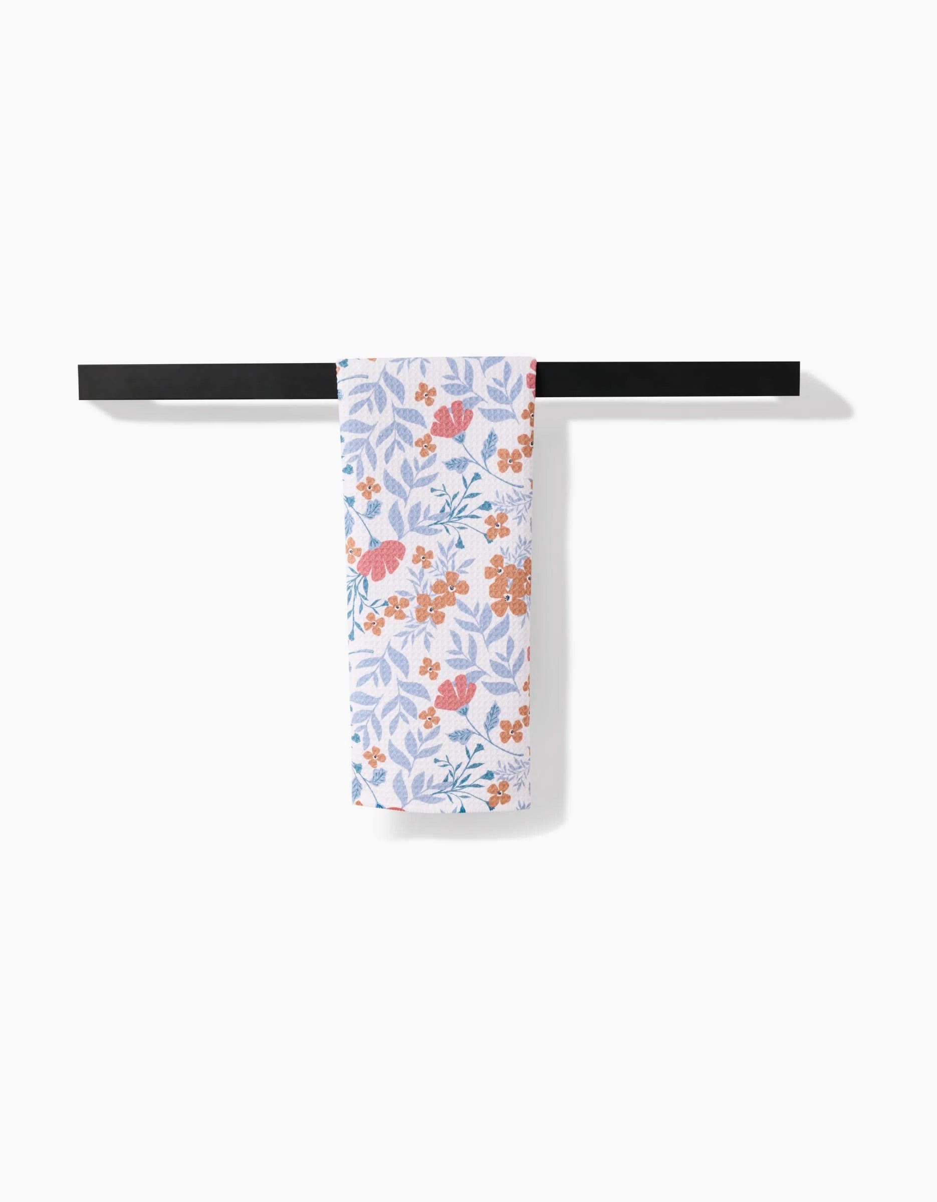 The Morning Blooms tea towel by Charlie Rowan Designs, featuring orange and blue floral and butterfly patterns on Geoweave fabric, hangs neatly on a sleek black rack against a plain white background.