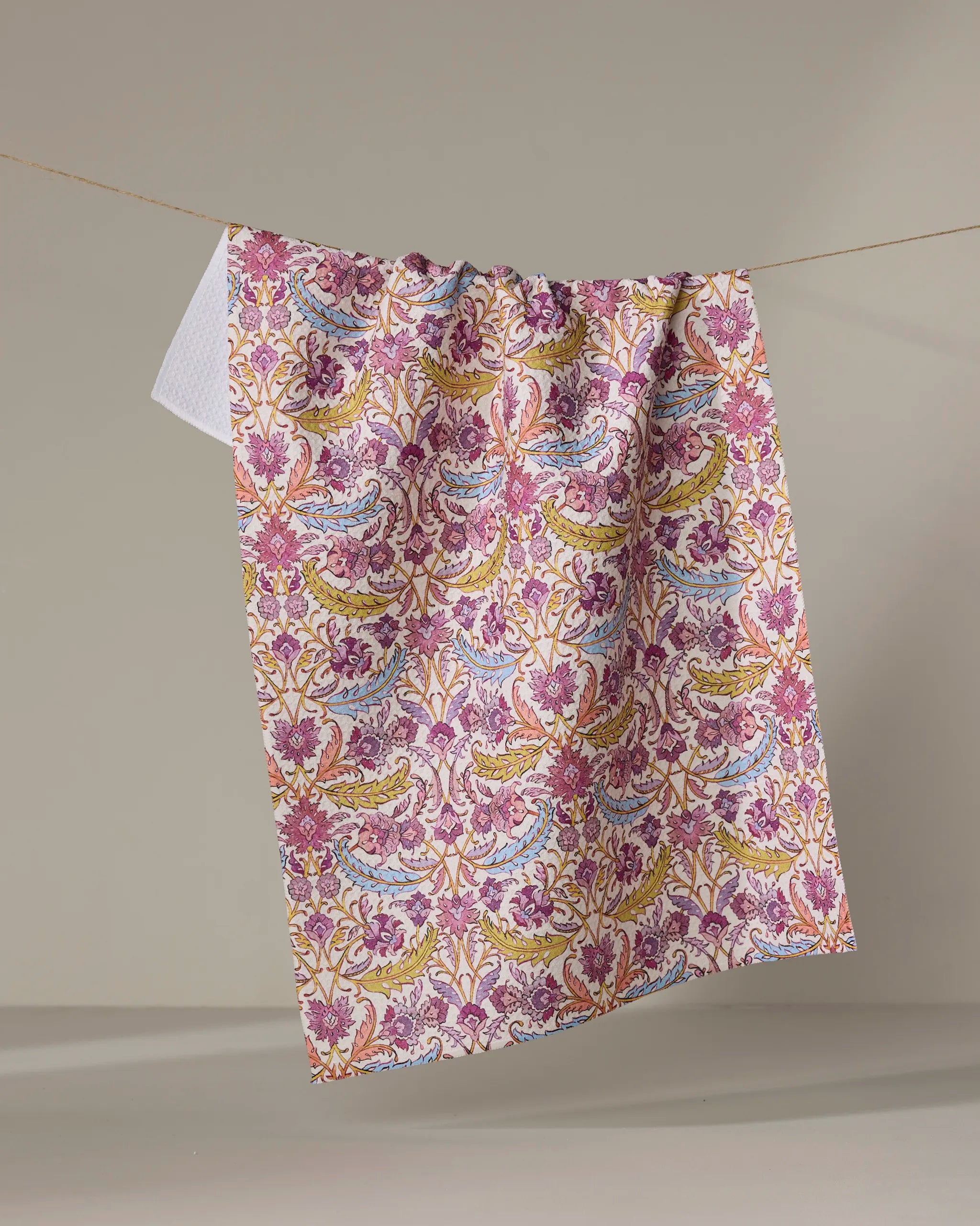 The “Morning Glory” tea towel by Maayan Alper-Swan, featuring colorful pink, yellow, green, and blue floral and leafy designs, hangs on a clothesline against a plain beige background.