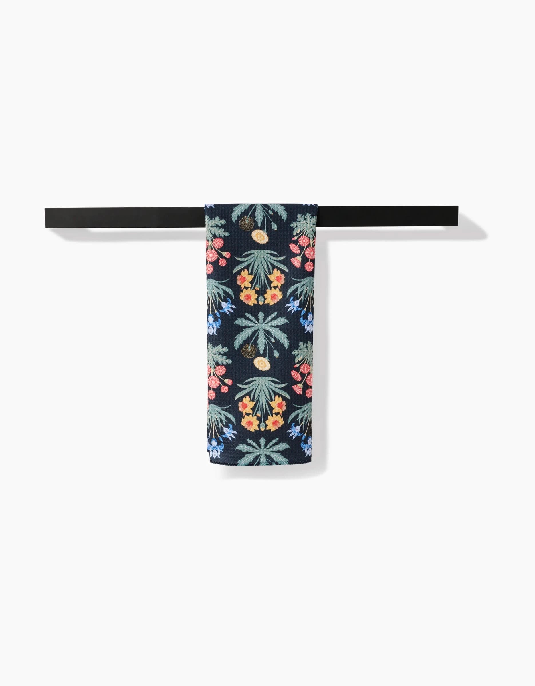 A black towel bar holds Danelys Sidron’s Morning Meadow dark blue tea towel, featuring a vibrant tropical floral pattern with palm trees, flowers, and fruit on a white background.